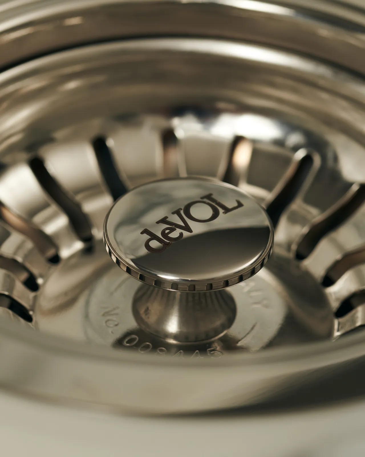 A close-up view of the central finial of a polished chrome basket waste strainer, there is a small 'deVOL' logo in the middle of the finial.
