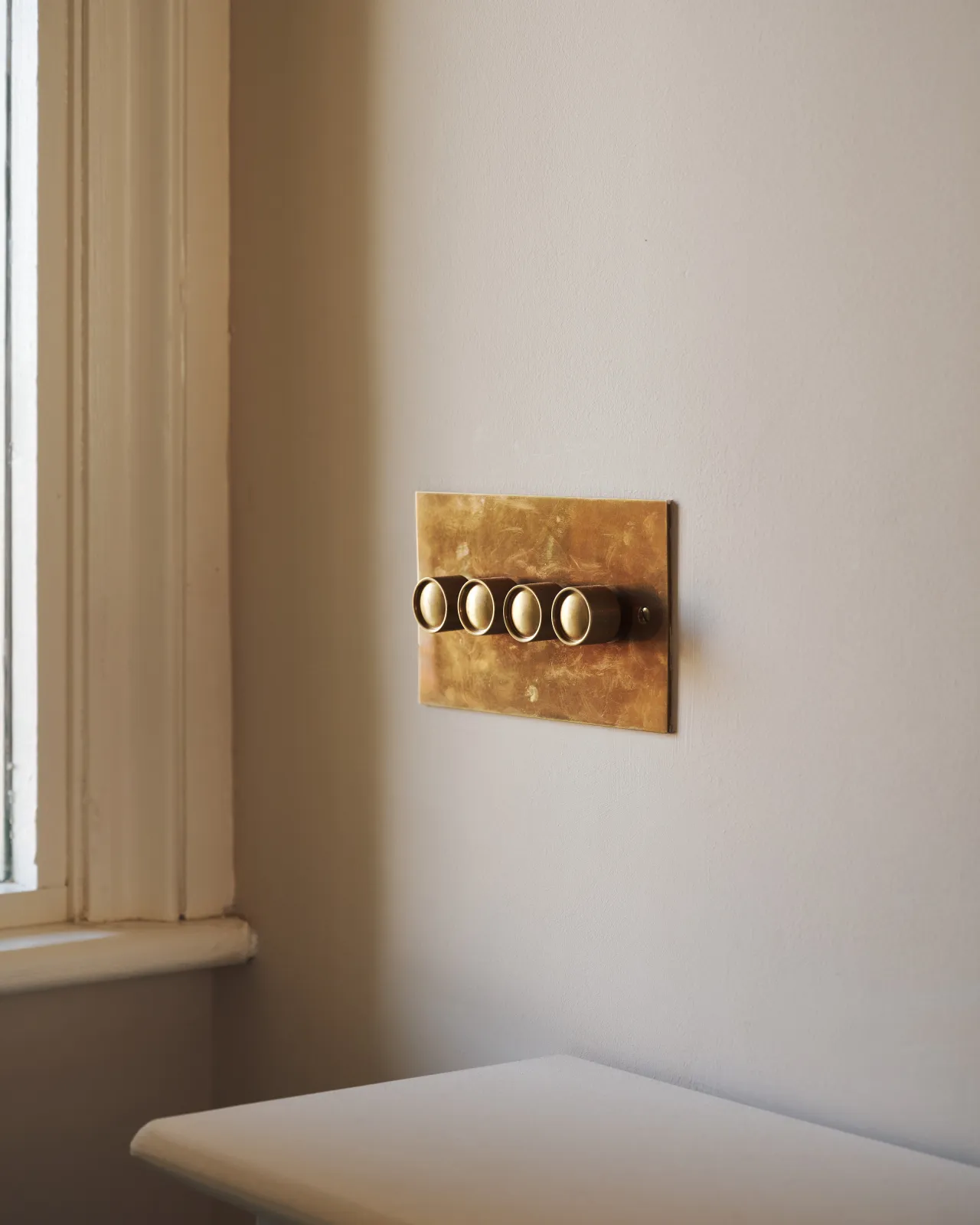 An oblique view of a 4 gang classic dimmer switch in a warm burnished brass finish on soft pink walls with light painted woodwork.