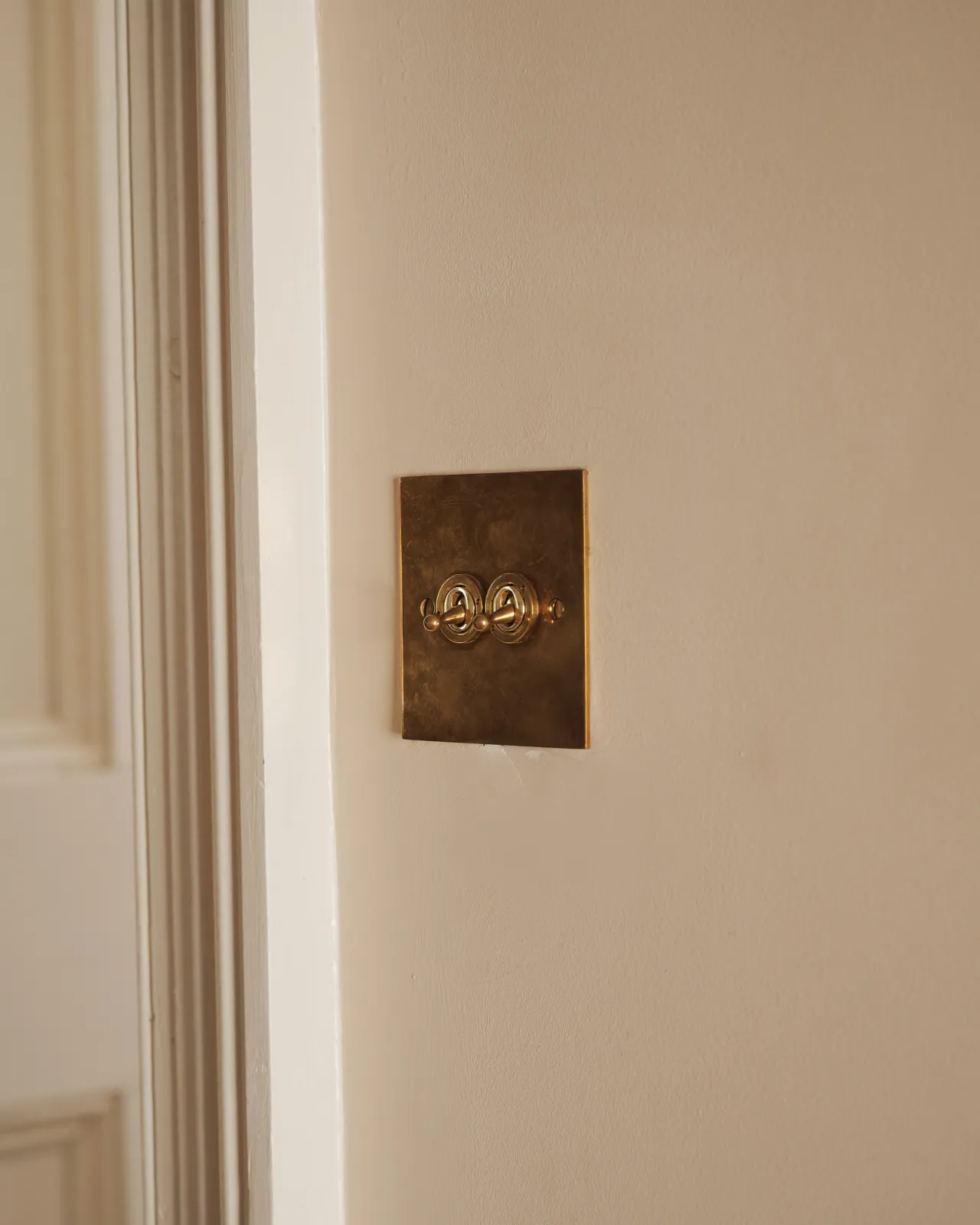 An oblique view of a 2 gang classic toggle switch in a warm burnished brass finish on soft pink walls with light painted woodwork.