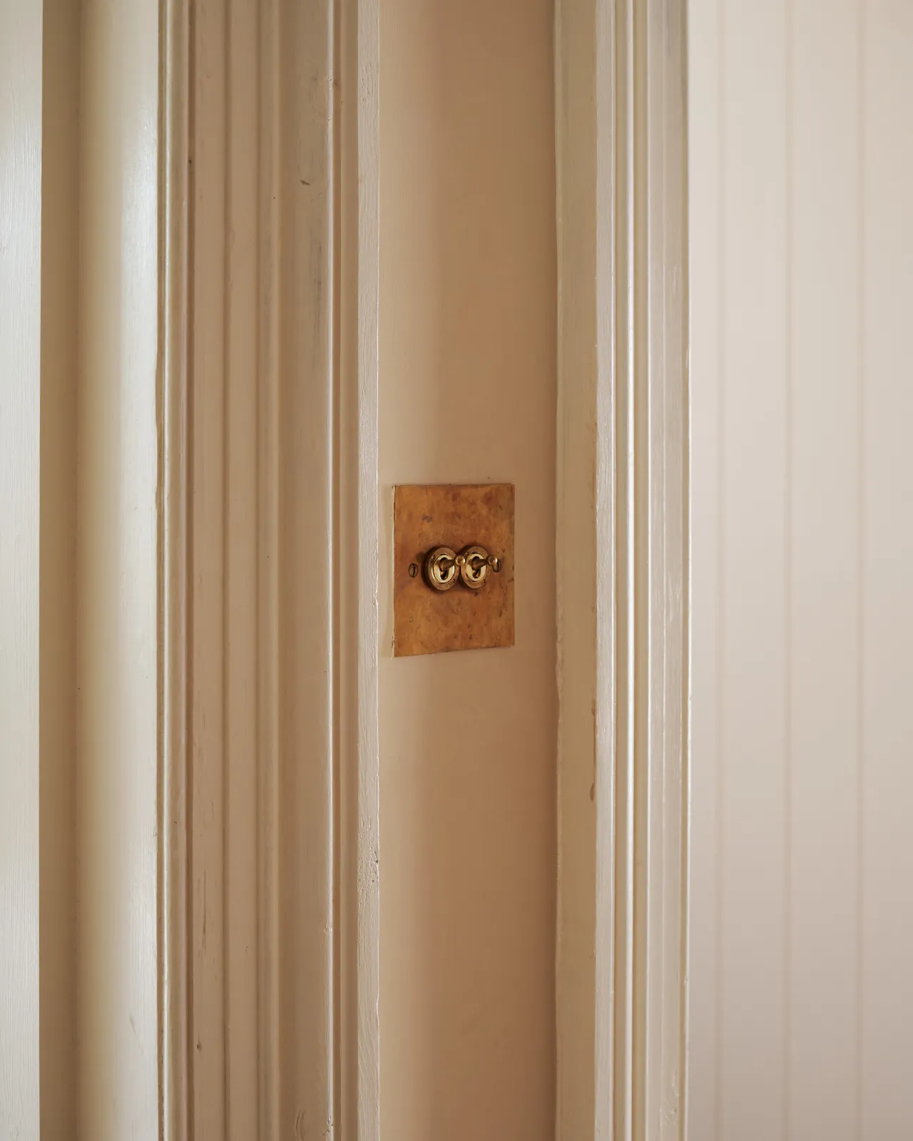 An oblique view of a 2 gang classic toggle switch in a warm burnished brass finish on soft pink walls between traditional light painted architrave.