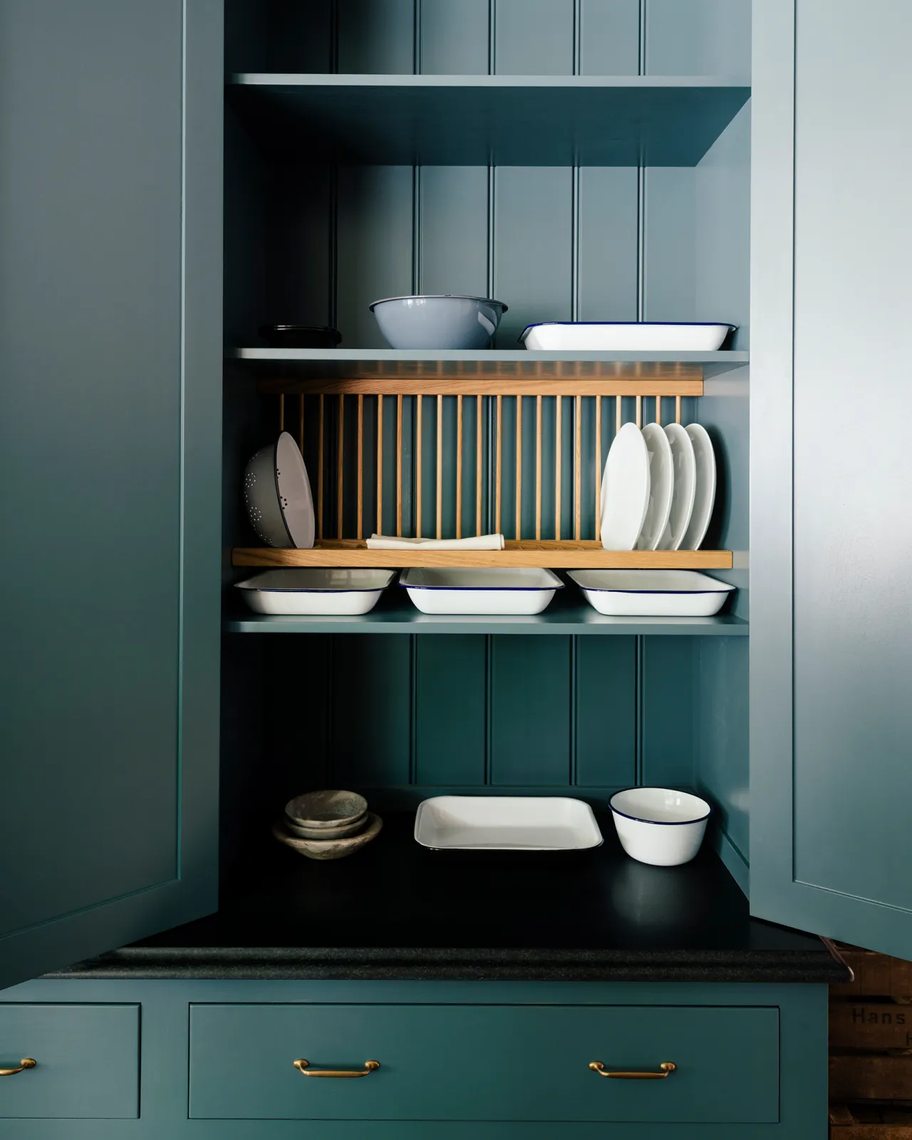 Looking into a Georgian-style pantry cupboard, painted inside and out in a green-tinged blue. Inside the cupboard there is wooden plate rack and a collection of white plates and enamel dishes.