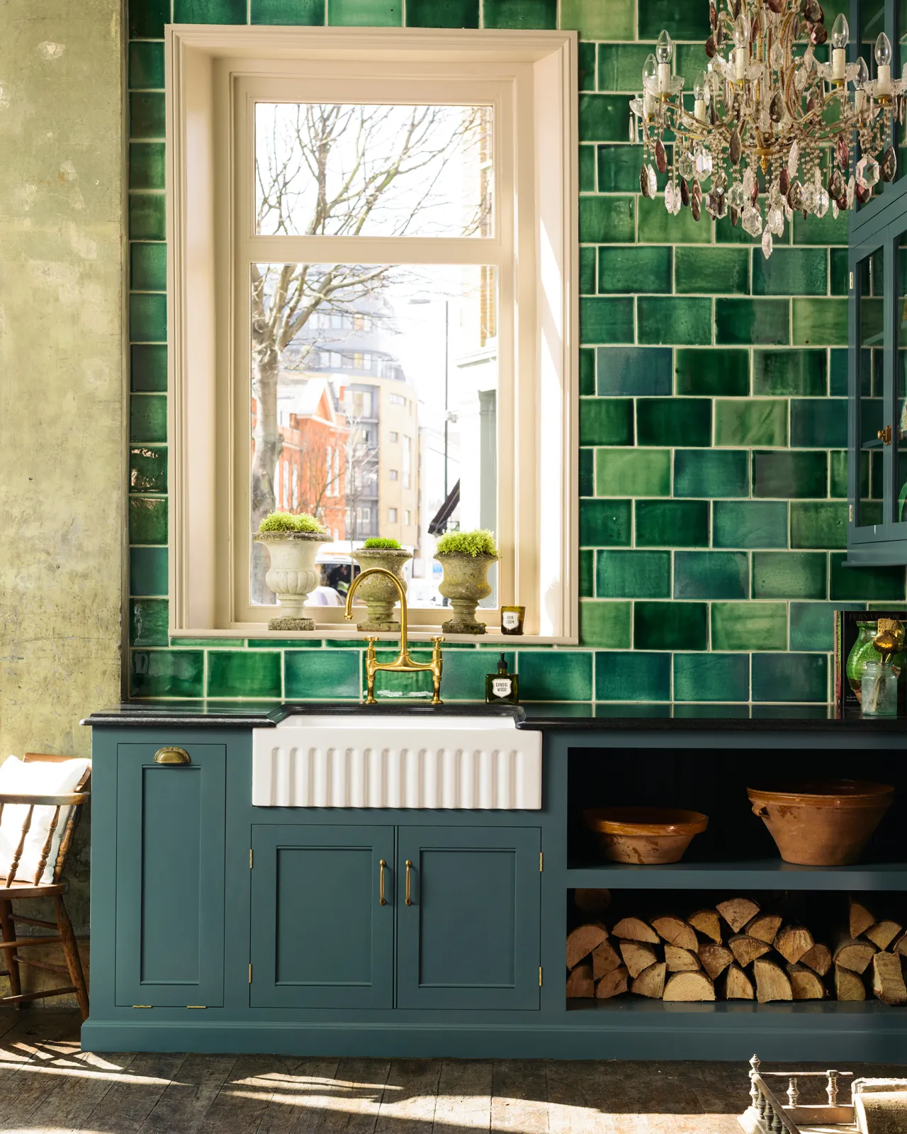 A straight-on view of a Georgian-style kitchen sink cabinet painted in a rich green-blue, with a black granite worktop, a wall of glossy emerald green tiles and an antique chandelier hanging high above.