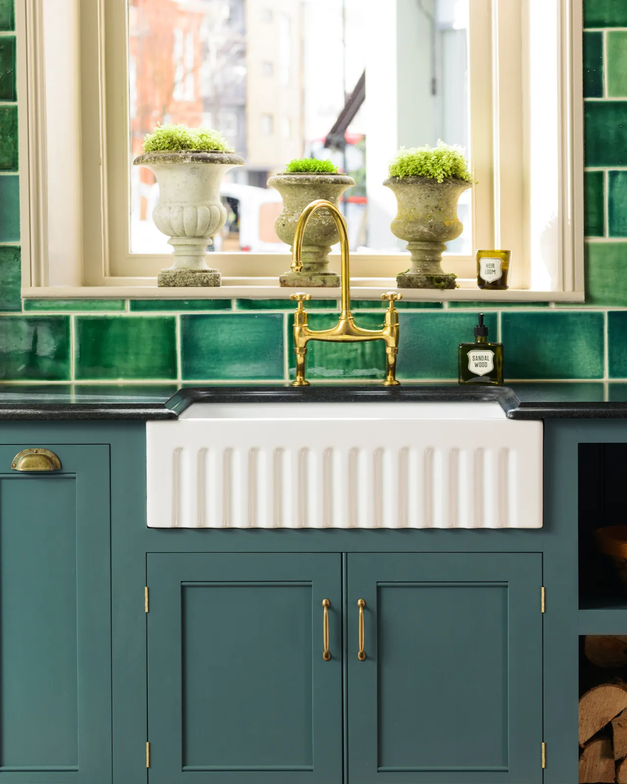 A close-up view of a Georgian-style kitchen sink cabinet painted in a rich green-blue, with a fluted Belfast sink, aged brass tap, black granite worktop, and a wall of glossy emerald green tiles behind.