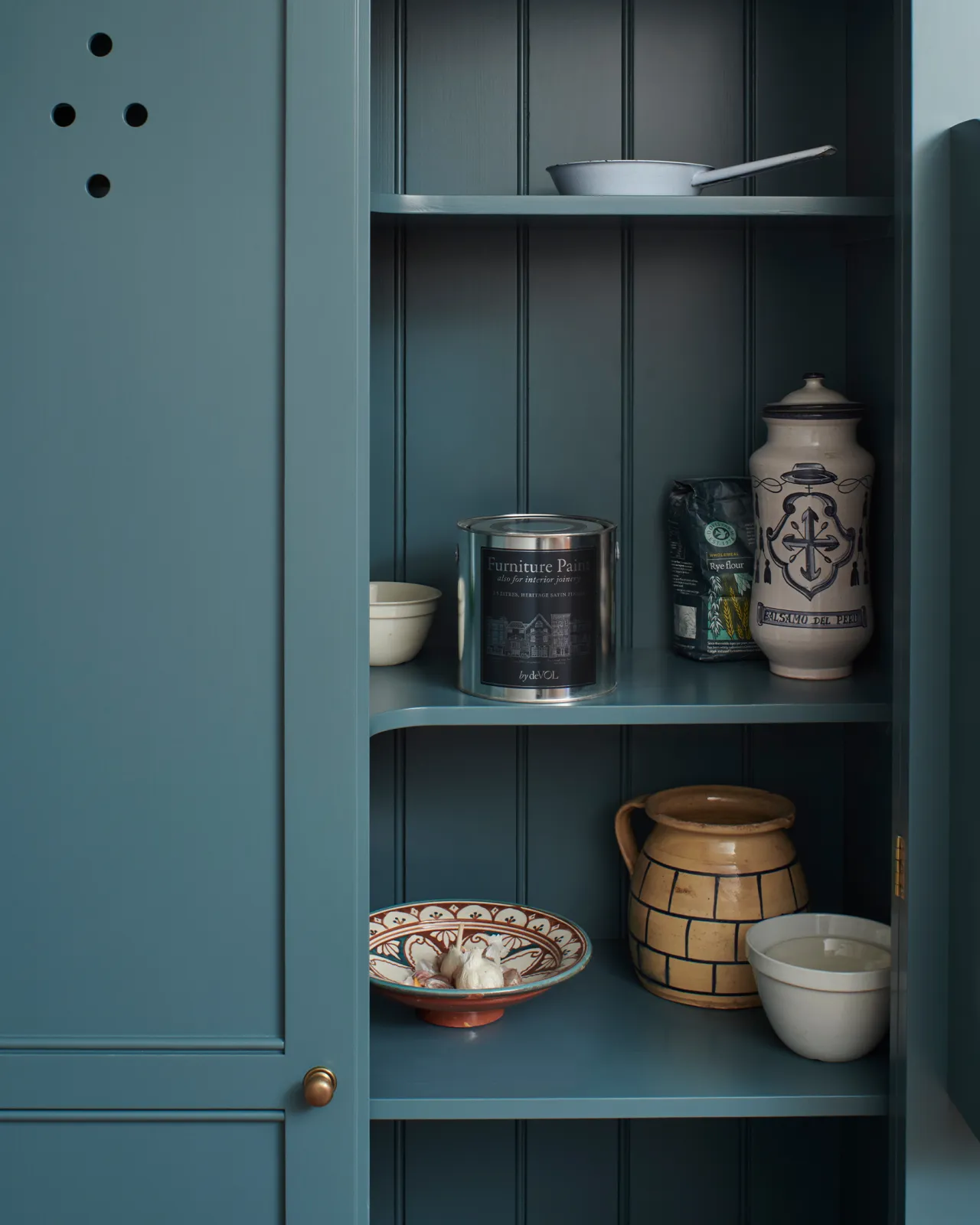 Looking into a Georgian-style pantry cupboard, painted inside and out in a green-tinged blue. Inside the cupboard there is a silver paint tin with a blue label and a collection of vintage crockery.