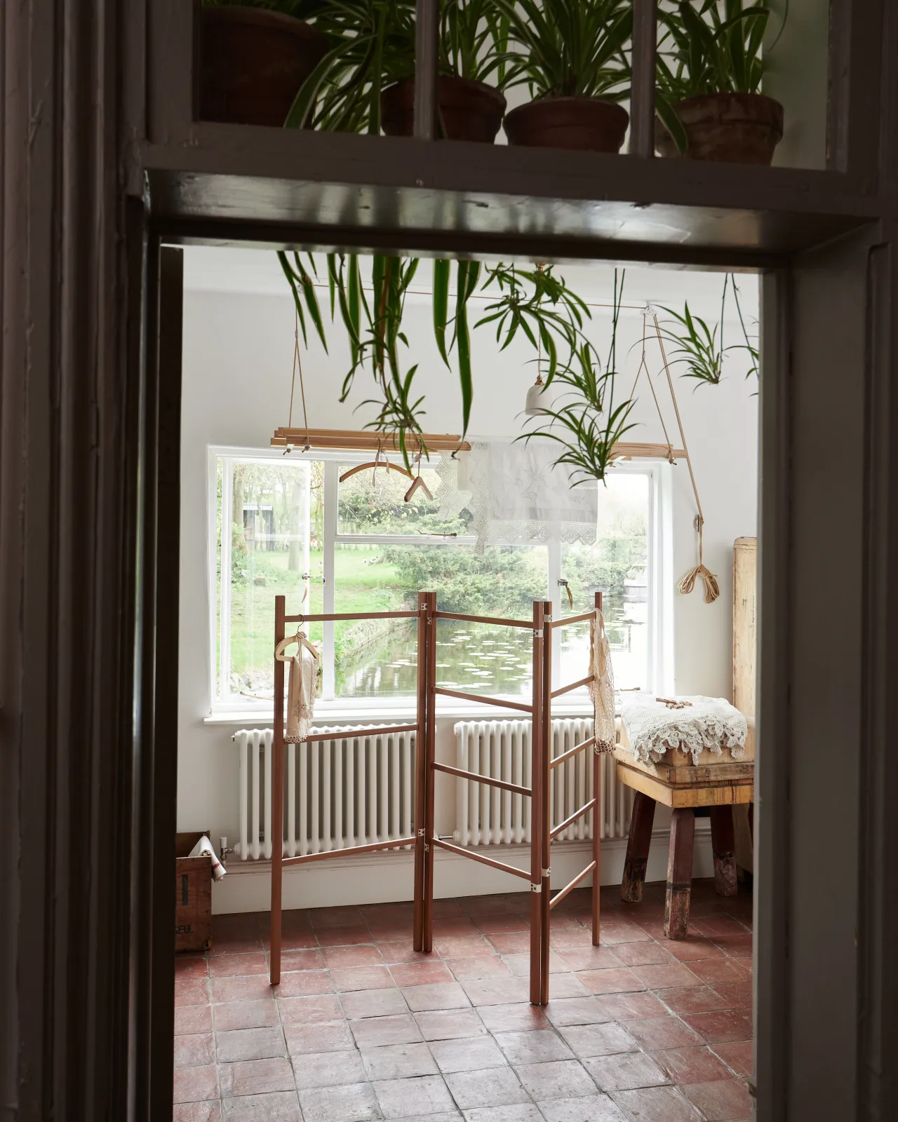 Looking through a doorway into a Mediterranean-inspired room with white walls and terracotta flooring and a wooden clothes horse.