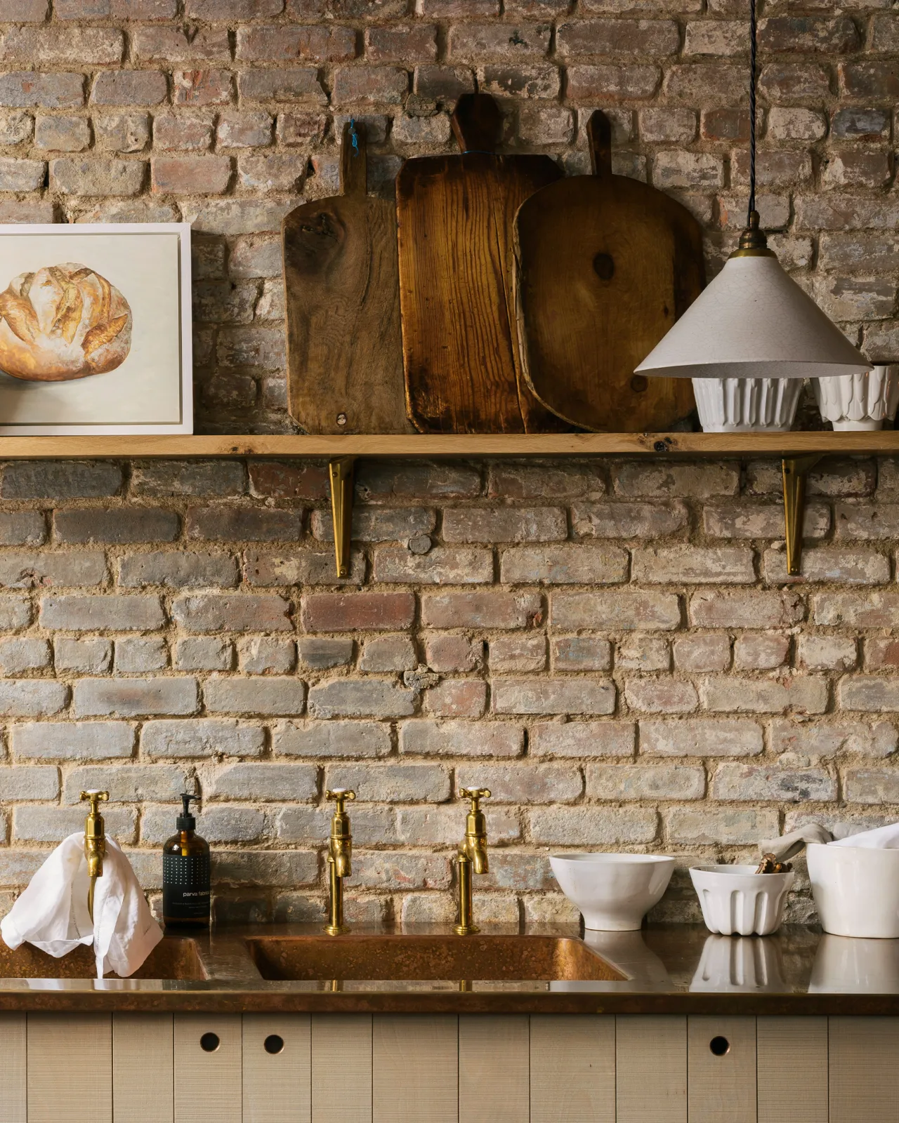 A straight-on view of a white glazed ceramic pendant light with delicate crazing hanging in front of a worn brick wall with an oak shelf.