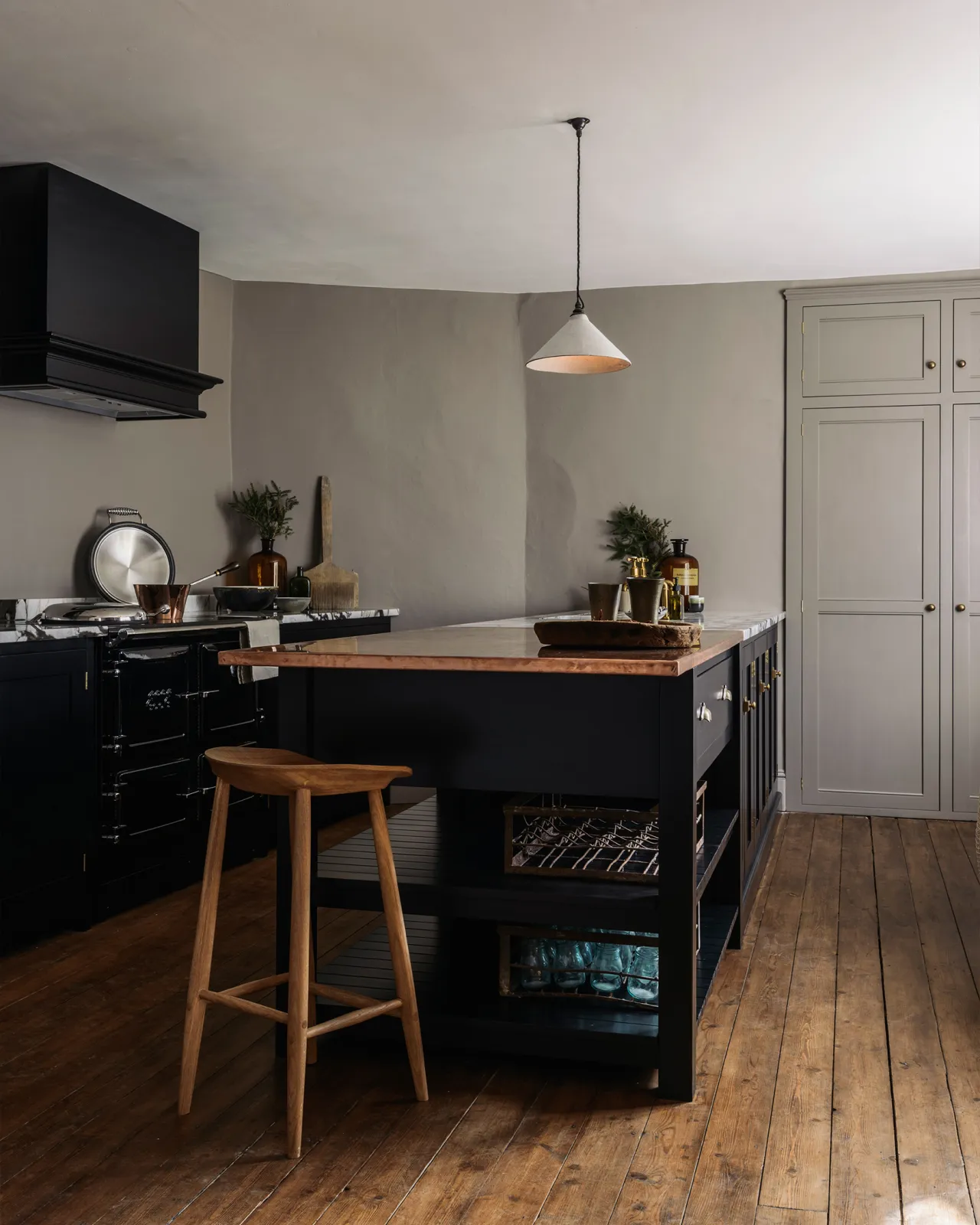 A white glazed ceramic pendant light with delicate crazing hanging in a Shaker-style kitchen with black painted cabinetry, wooden floorboards and grey walls.