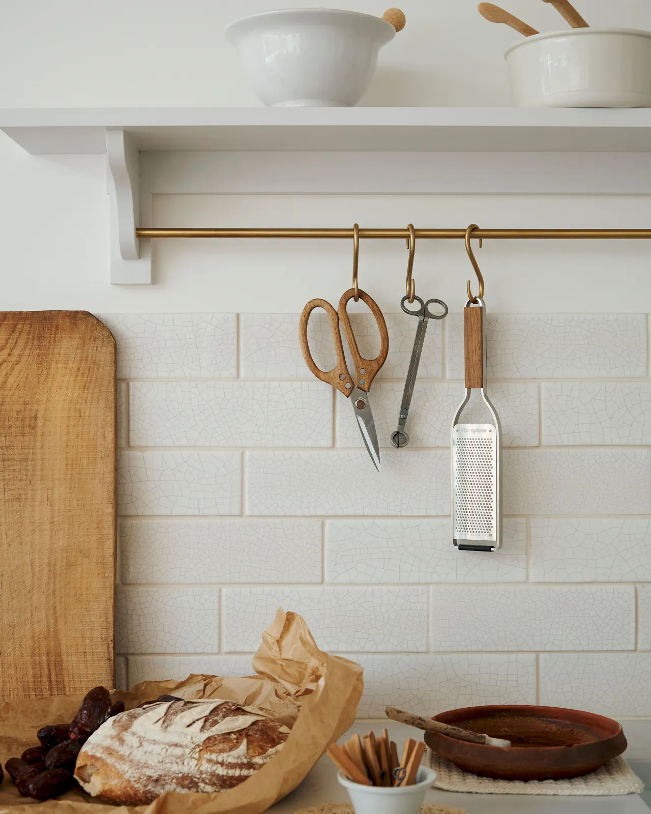 A close-up view of an assortment of kitchen utensils hanging from a brass rail in front of white metro wall tiles with delicate crazing.