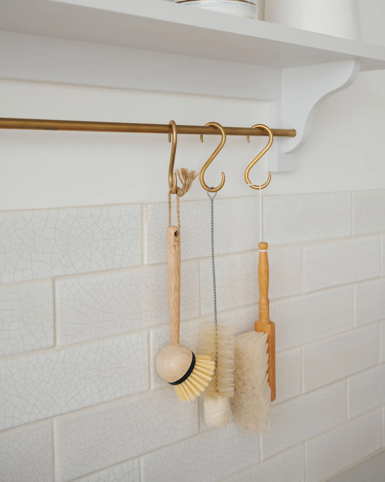 An oblique close-up view of an assortment of kitchen utensils hanging from a brass rail in front of white metro wall tiles with delicate crazing.