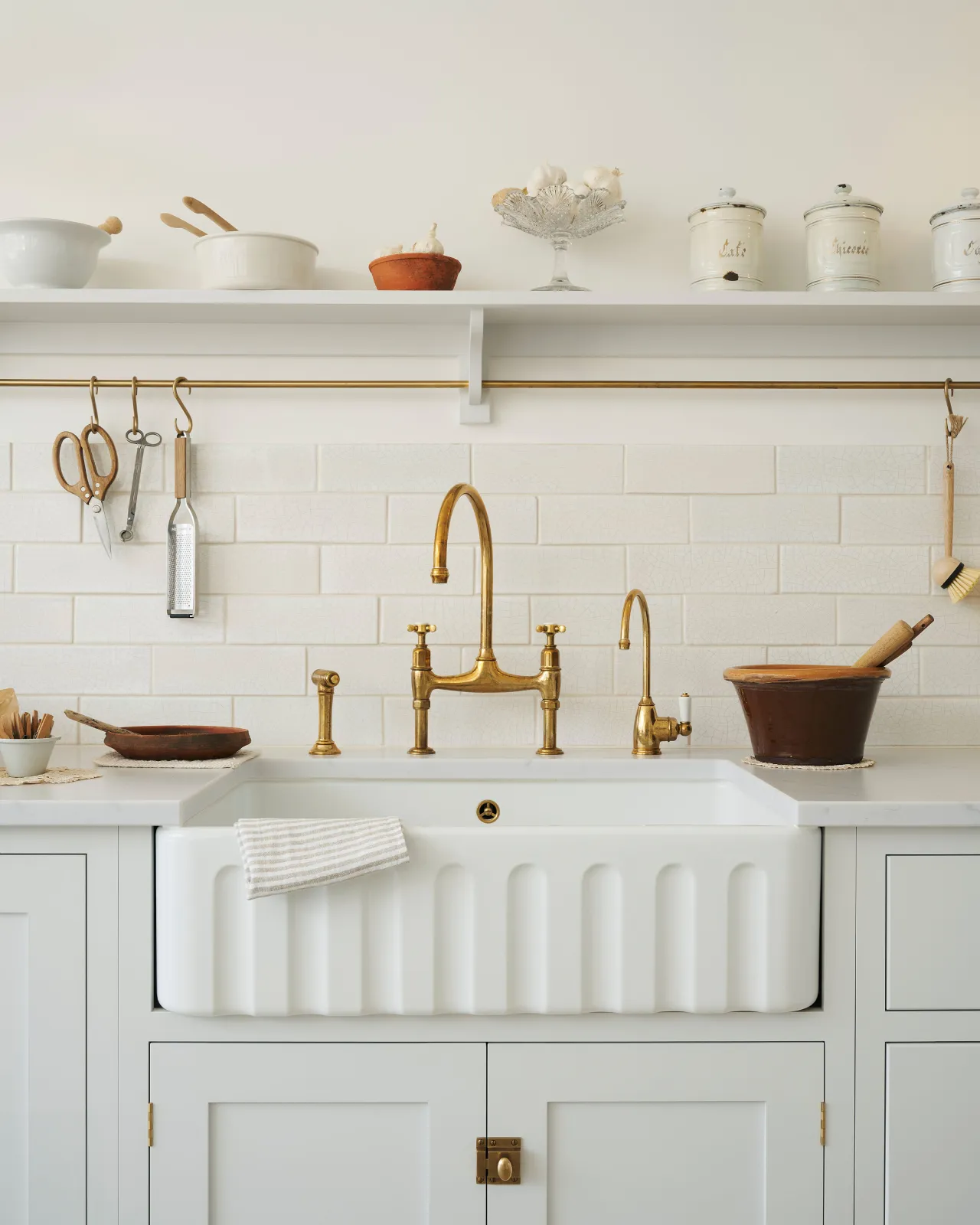 A close-up view of a fluted Belfast sink with white painted Shaker-style cupboard and an aged brass mixer tap. A simple shelf with a brass hanging rail and a splashback of white metro wall tiles with delicate crazing sits behind the tap.