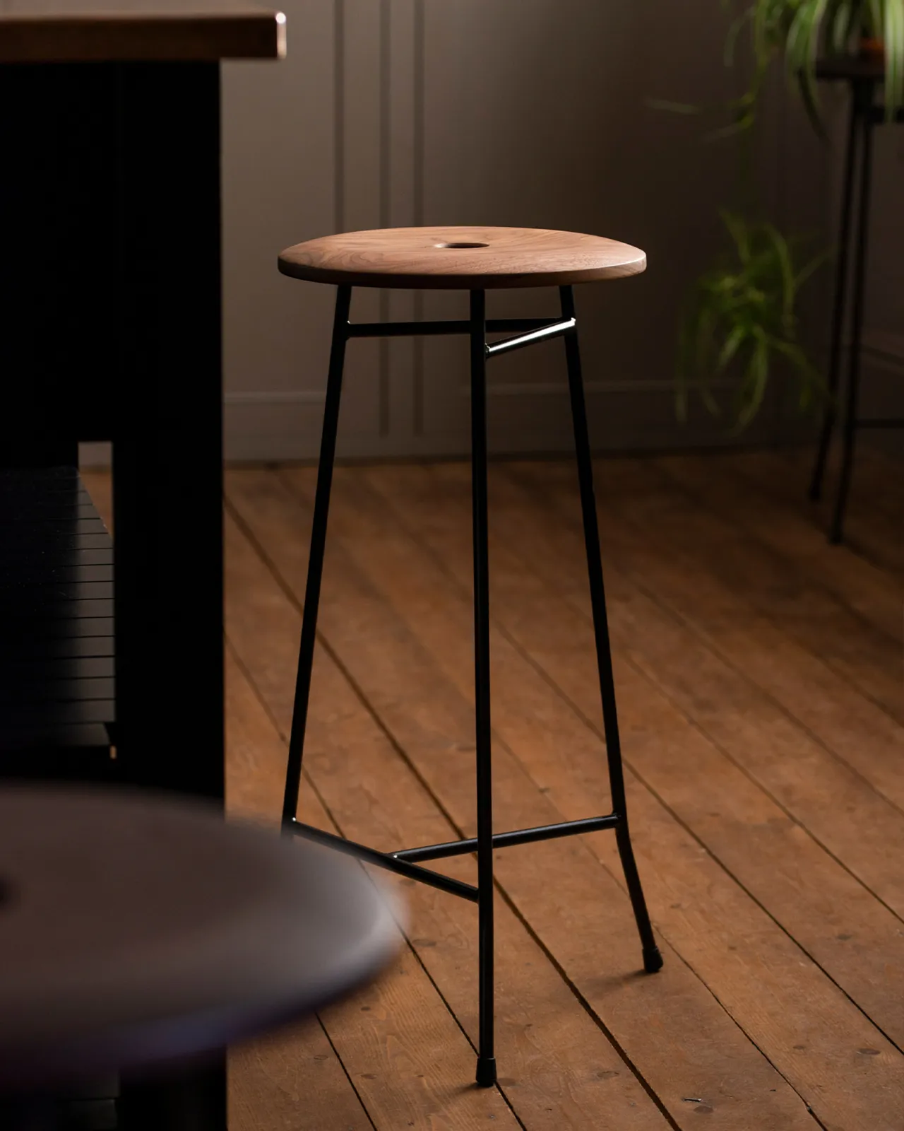 A mid-century-style three-legged bar stool with a thin dark metal frame and a round walnut seat top with a central hole and a natural oiled finish.