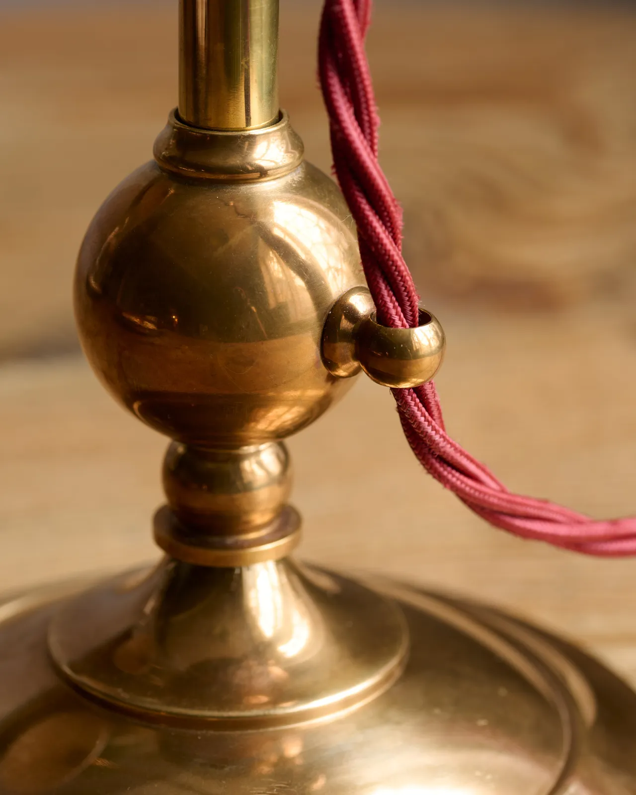 A close-up view of a brass bass finial of a Victorian-inspired desk lamp with a red twisted fabric flex cord running through a brass loop.