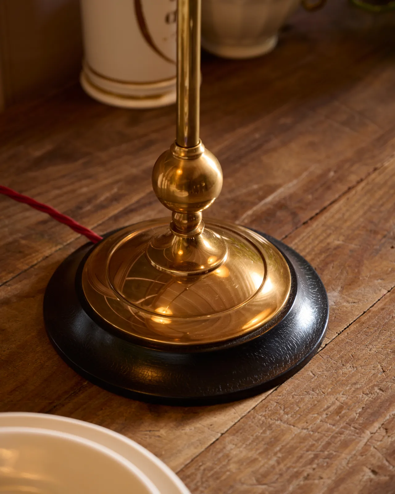 A close-up view of a solid brass desk lamp foot with a dark hardwood base and a red twisted fabric flex cord leading out from one side.