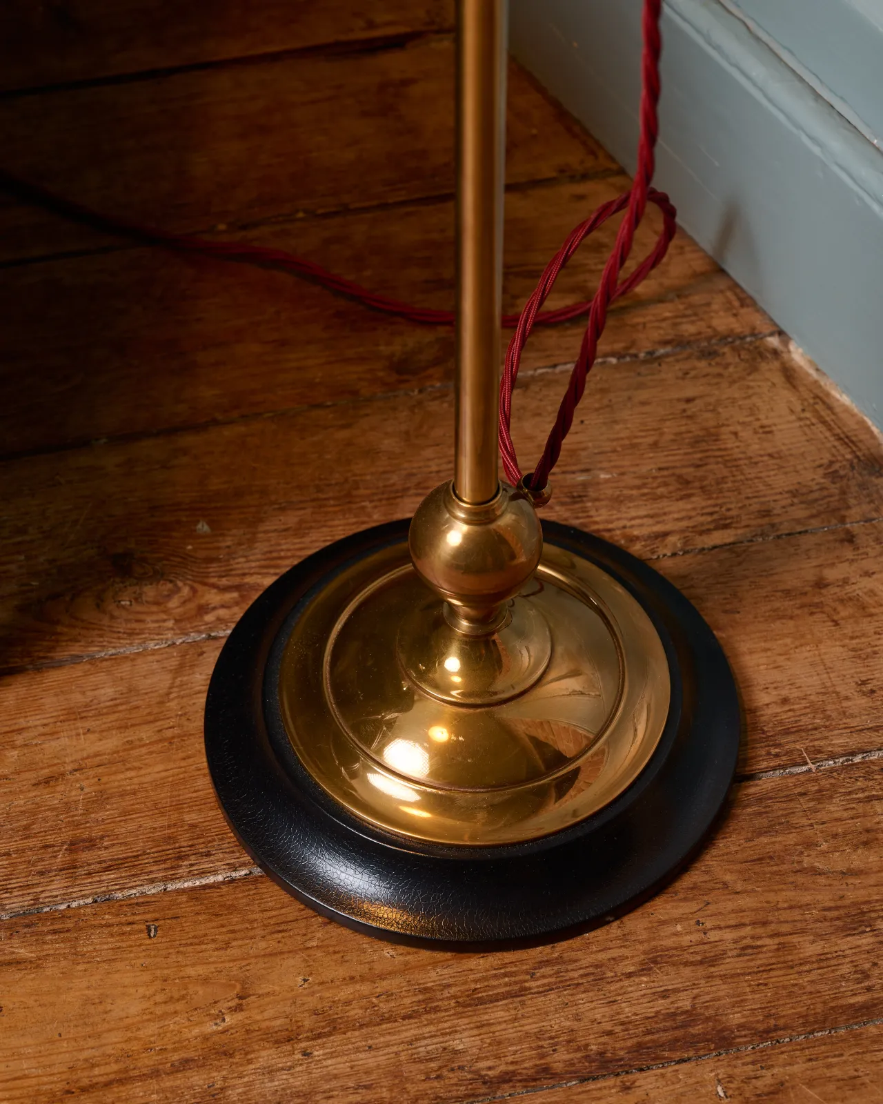 A close-up view of a solid brass floor lamp foot with a dark hardwood base and a red twisted fabric flex cord leading out from one side.