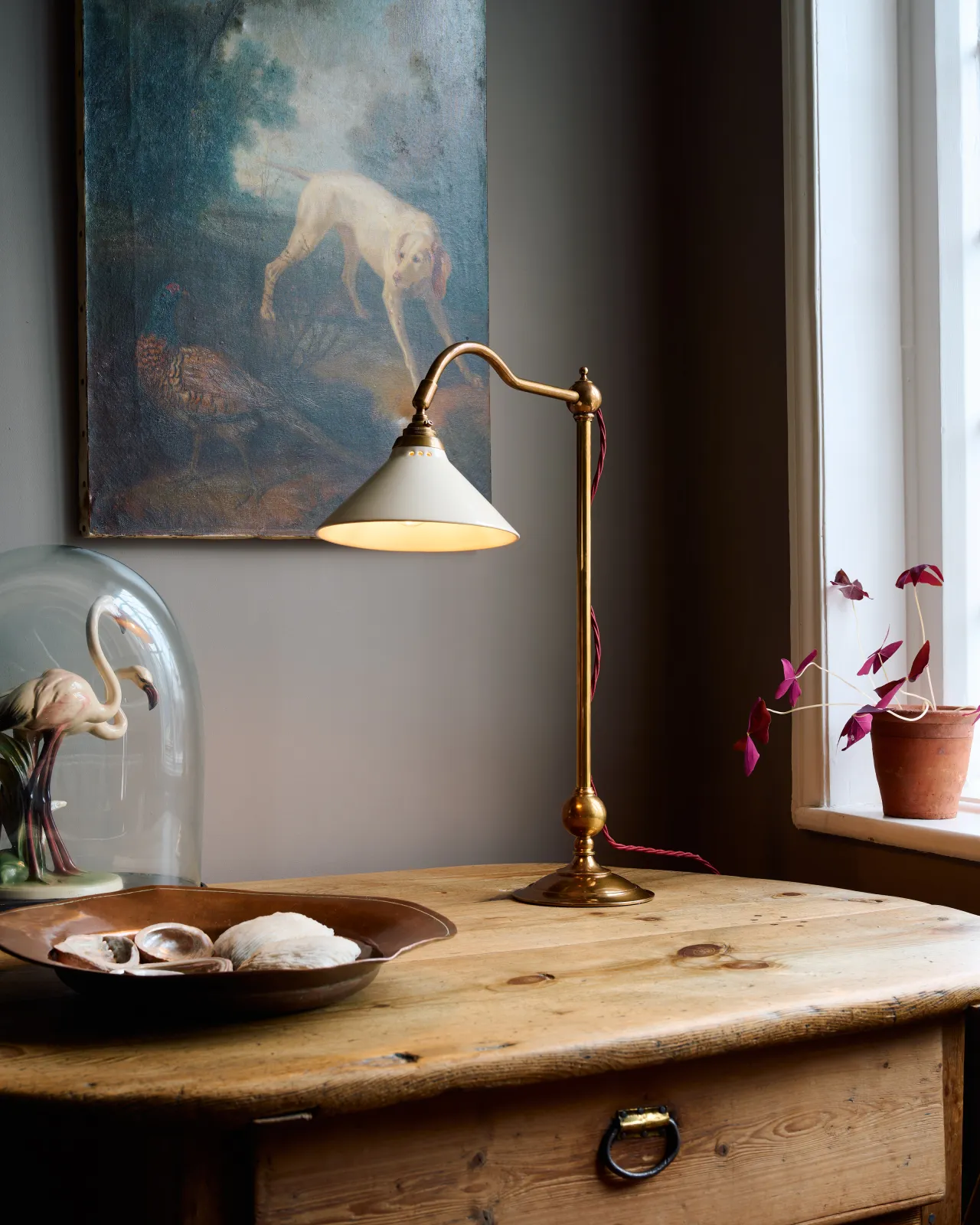 An oblique view of an antique table with a Victorian-inspired desk lamp with a solid brass foot, stand, curved arm and a small cream glazed ceramic light shade. A red twisted fabric flex cord can be seen coming from the top finial.
