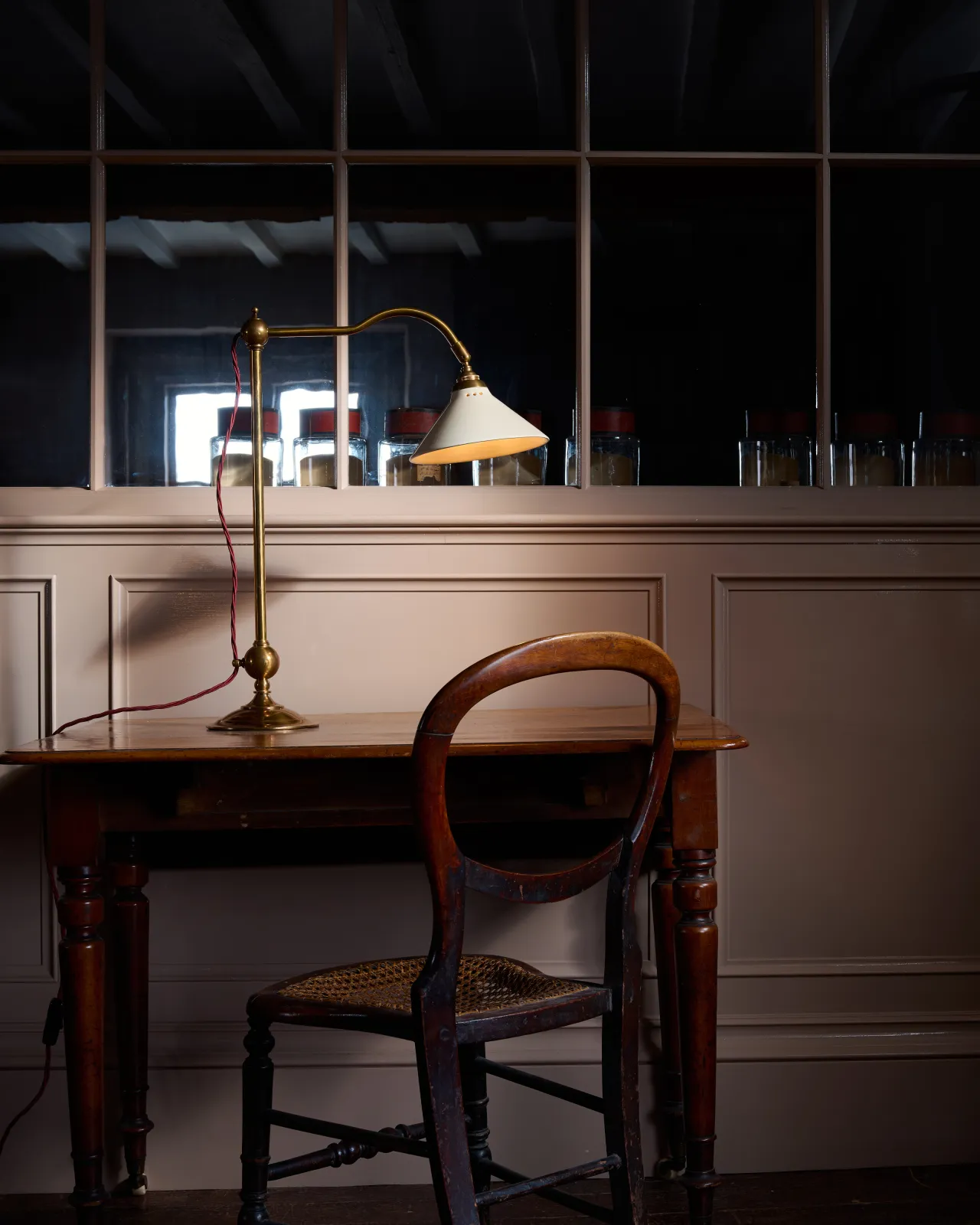 A straight-on view of an antique table in front of a pink painted glazed partition wall. On the table is a Victorian-inspired desk lamp with a solid brass foot, stand, curved arm and a small cream glazed ceramic light shade.