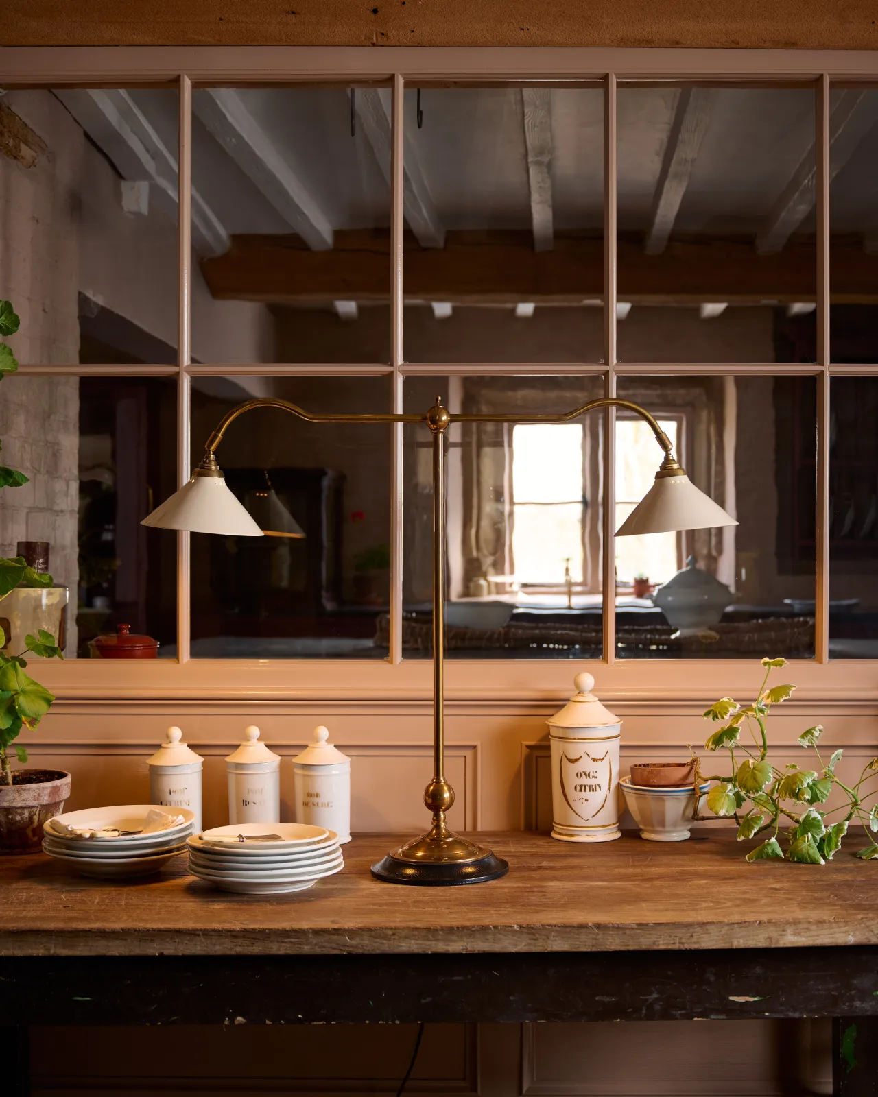 A straight-on view of a Victorian-inspired desk lamp with a solid brass foot, stand, two curved arms and two small cream glazed ceramic light shades on a table in front of a glazed partition.