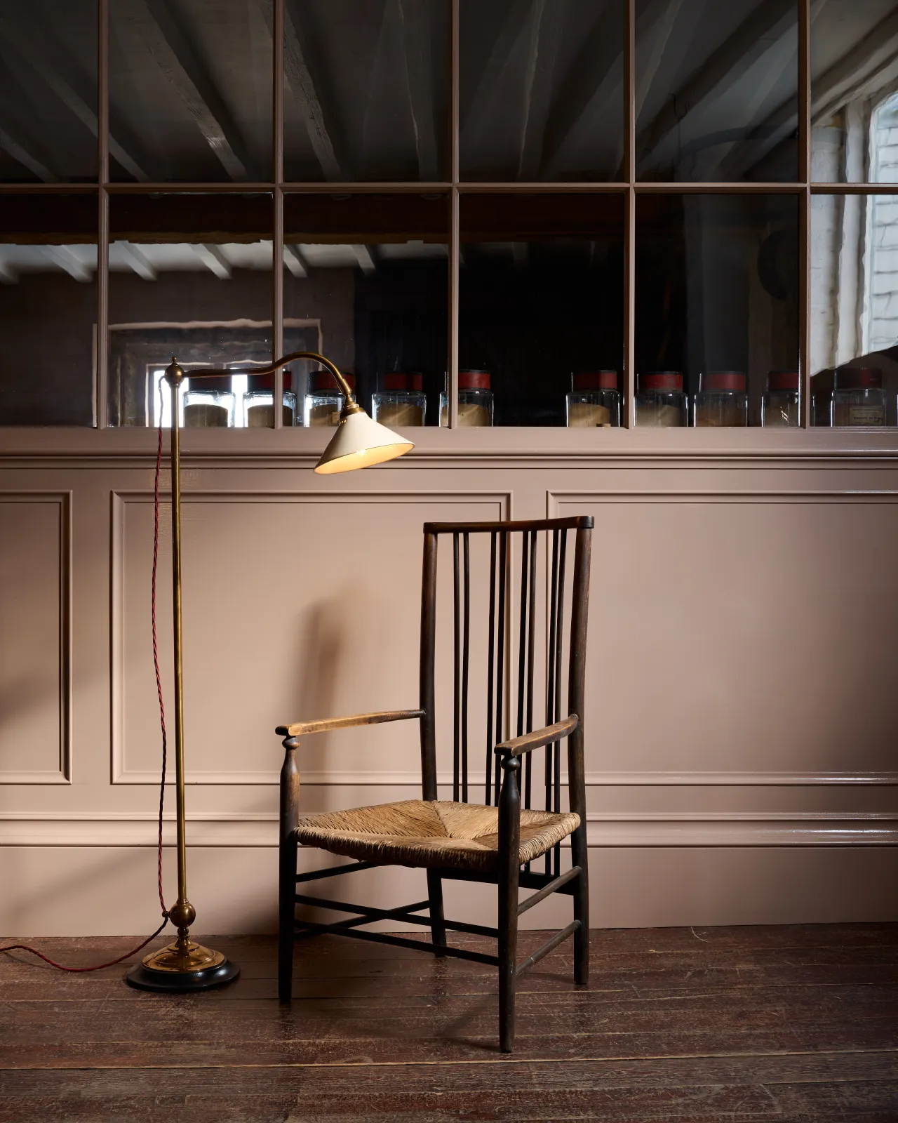 A straight-on view of a Victorian-inspired floor lamp with a solid brass foot, stand, curved arm, dark hardwood base and a small cream glazed ceramic light shade, beside an antique wooden chair, in front of a pink painted glazed partition wall.