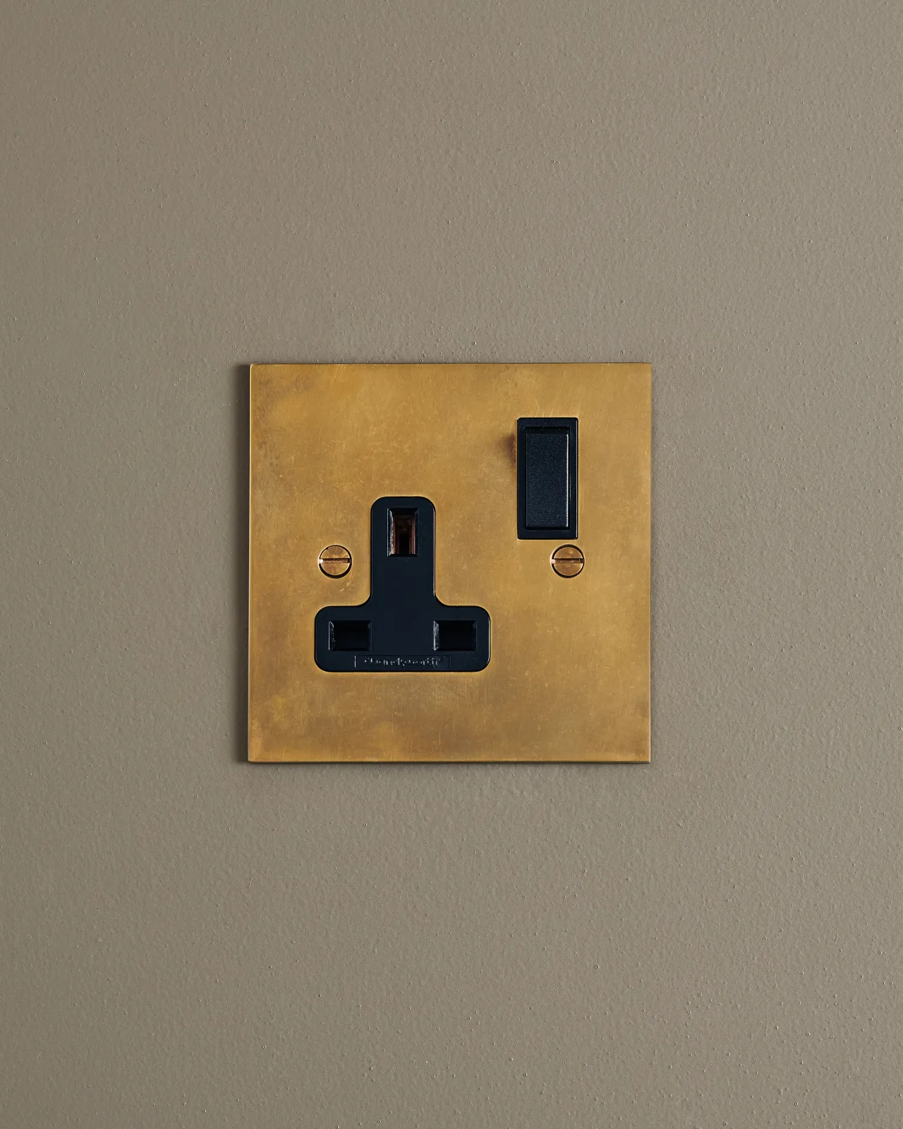 A straight-on view of a classic 13 amp single plug socket in a warm burnished brass finish with black plastic switches.