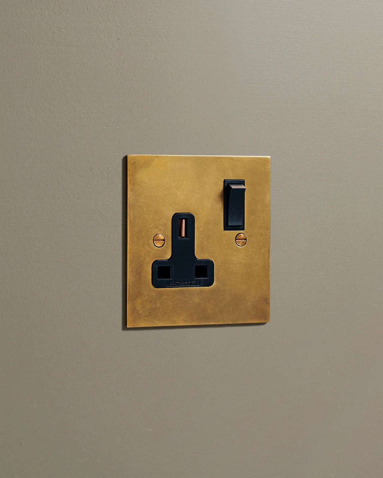 An oblique view of a classic 13 amp single plug socket in a warm burnished brass finish with black plastic switches.