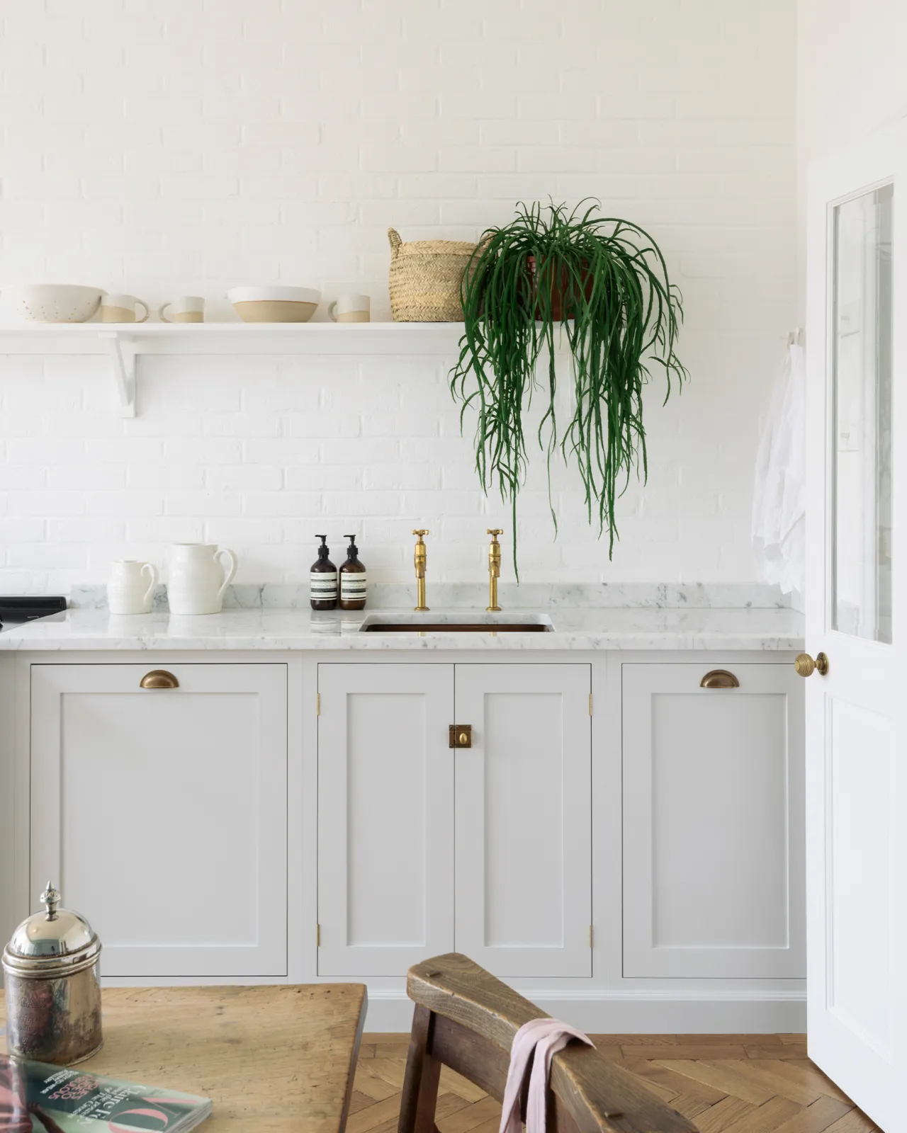 A straight-on view of a simple Shaker-style kitchen with off-white painted cupboards, white walls and a shelf with a very lush hanging plant.