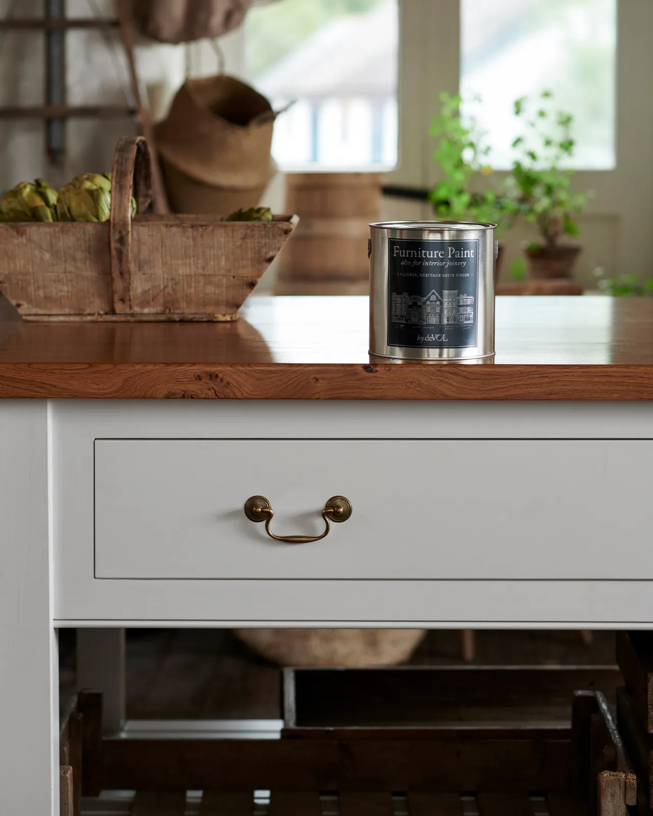 A straight-on view of an off-white painted prep table with a rustic oak worktop, wide drawers with Georgian-style pull handles. There is a silver paint tin with a blue label on the worktop.