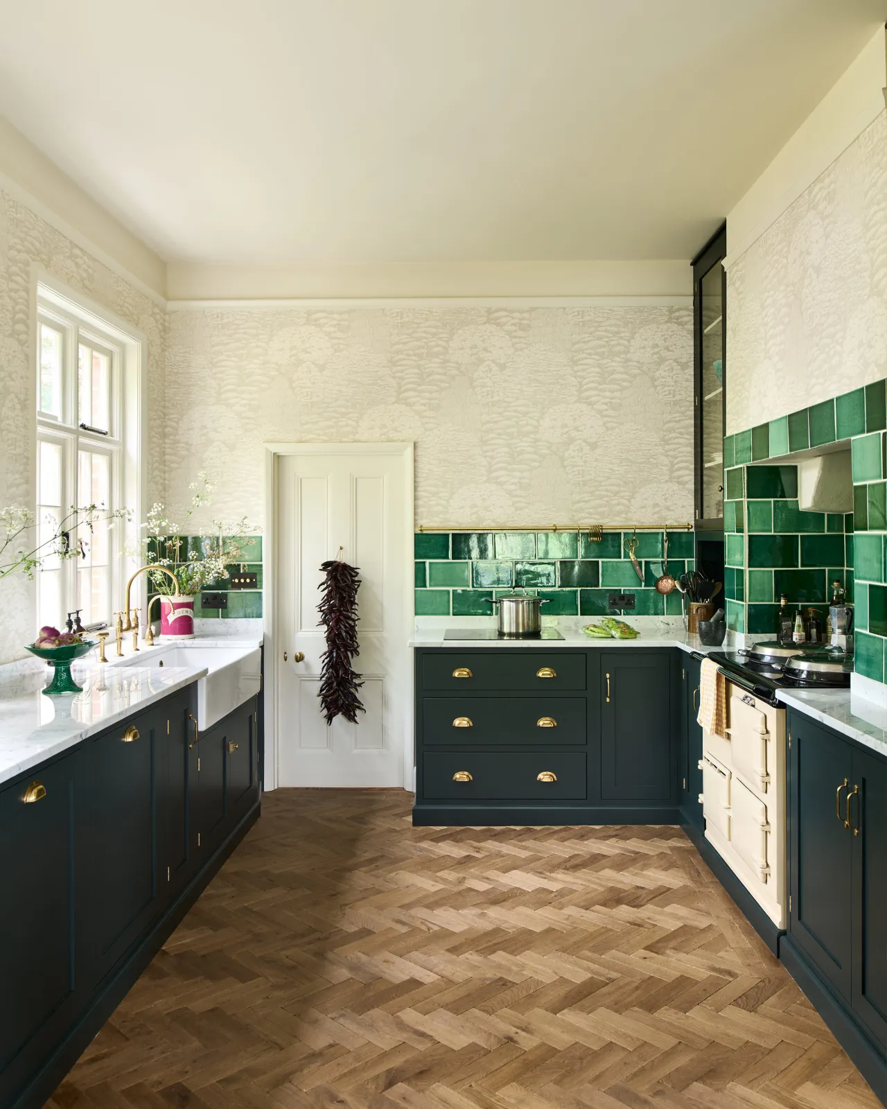 Looking into a classic kitchen with herringbone wood floor, dark green painted Shaker-style cupboards and creamy patterned wallpaper. A splashback of glossy emerald green wall tiles borders the kitchen worktop around the room.