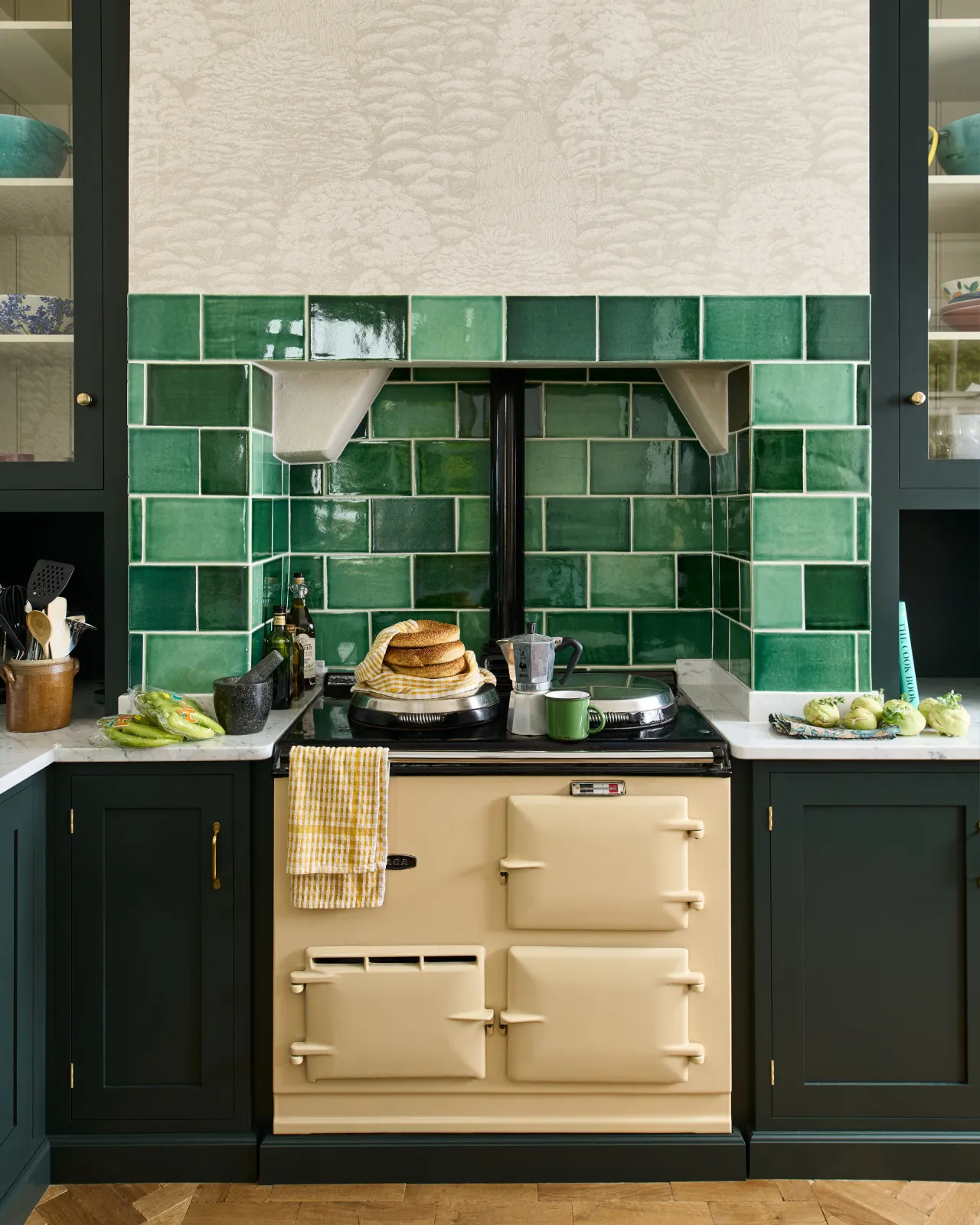 A straight-on view of a creamy Aga cooker with dark green kitchen cupboards either side and a beautifully glossy splashback of emerald green wall tiles.