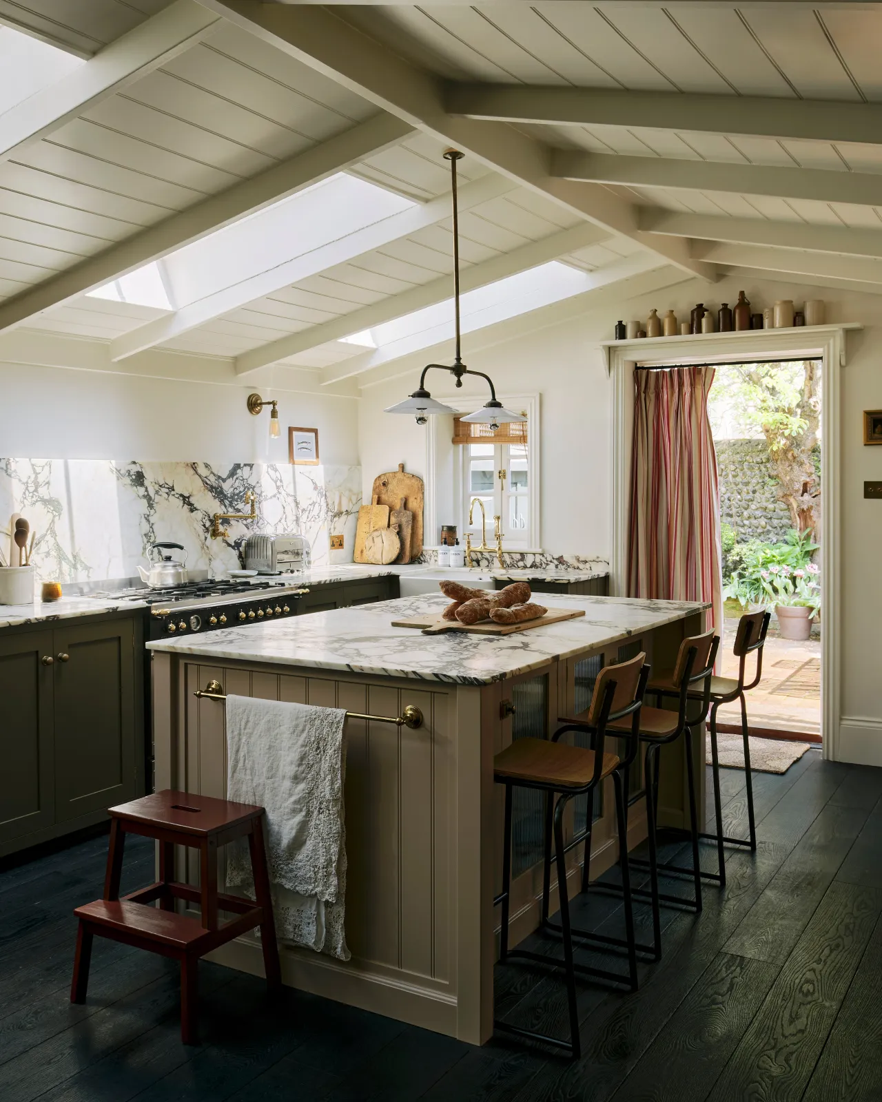 A grey painted Shaker-style kitchen with a white painted wooden pitched ceiling and an island with two industrial bar stools with oak veneer plywood seat pans and backrests.