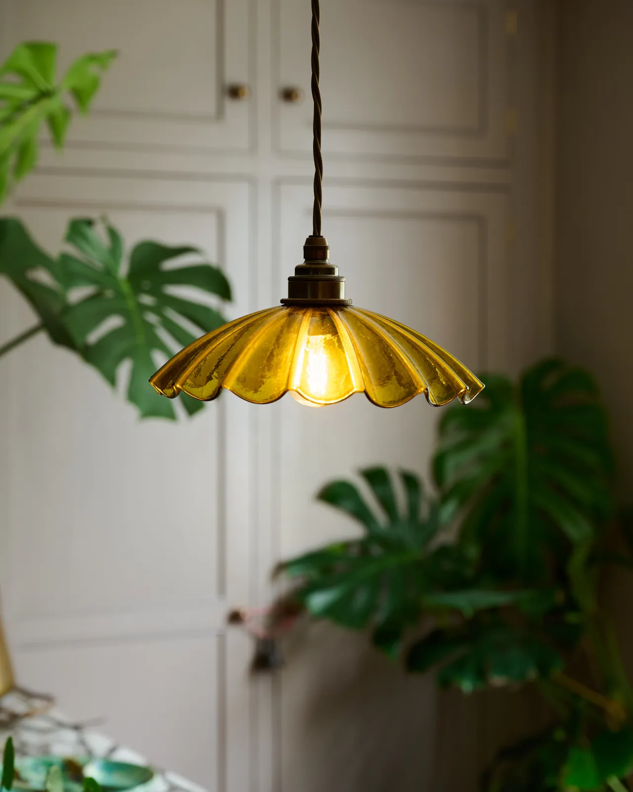 A small frilly chartreuse glass pendant light with brass fittings and brown twisted fabric flex cord. The light is on and shining through the tranlucent glass.