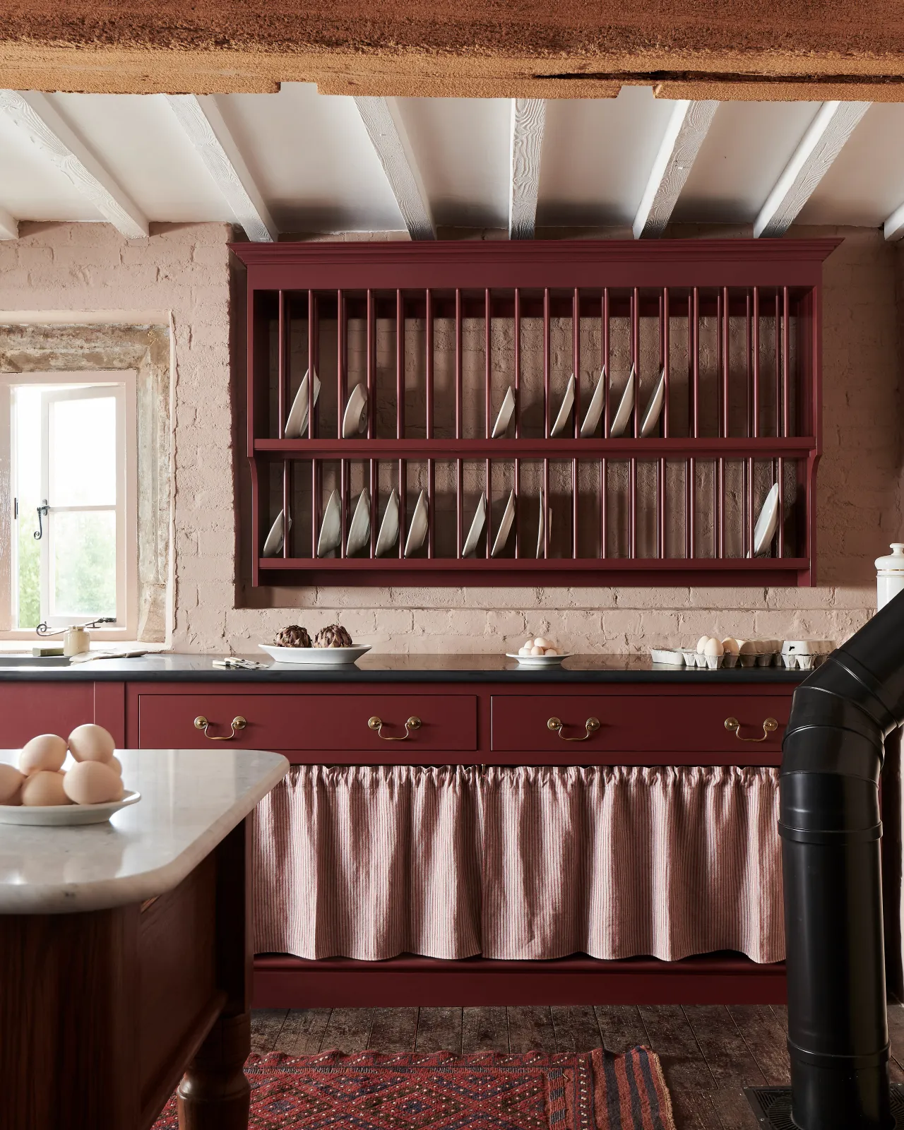A straight-on view of a Victorian-inspired kitchen with a traditional wall mounted plate rack hanging over a drawer cupboard with open storage behind a striped curtain. The drawers are fitted with Georgian-style swing handles in a burnished 'Bella' brass finish.