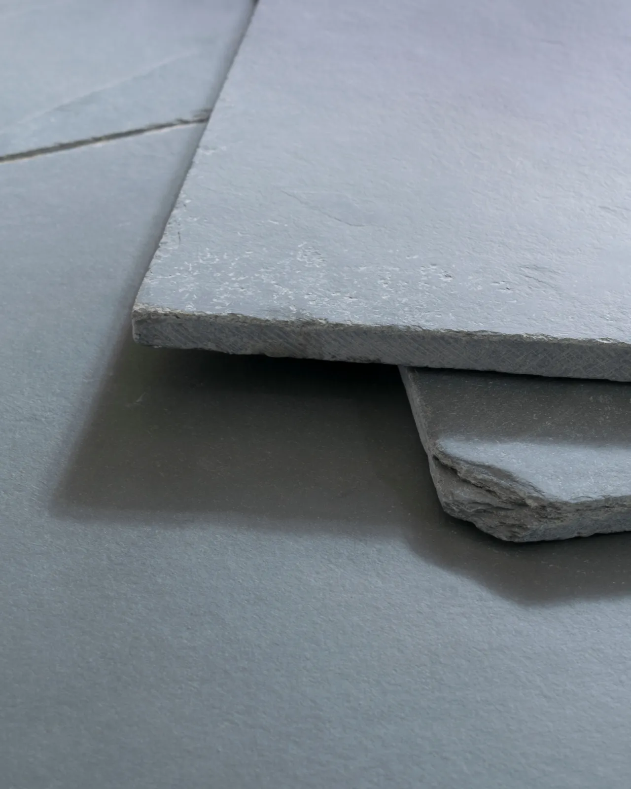 A close-up oblique view of a grey tumbled slate floor, with two loose tiles stacked on top.