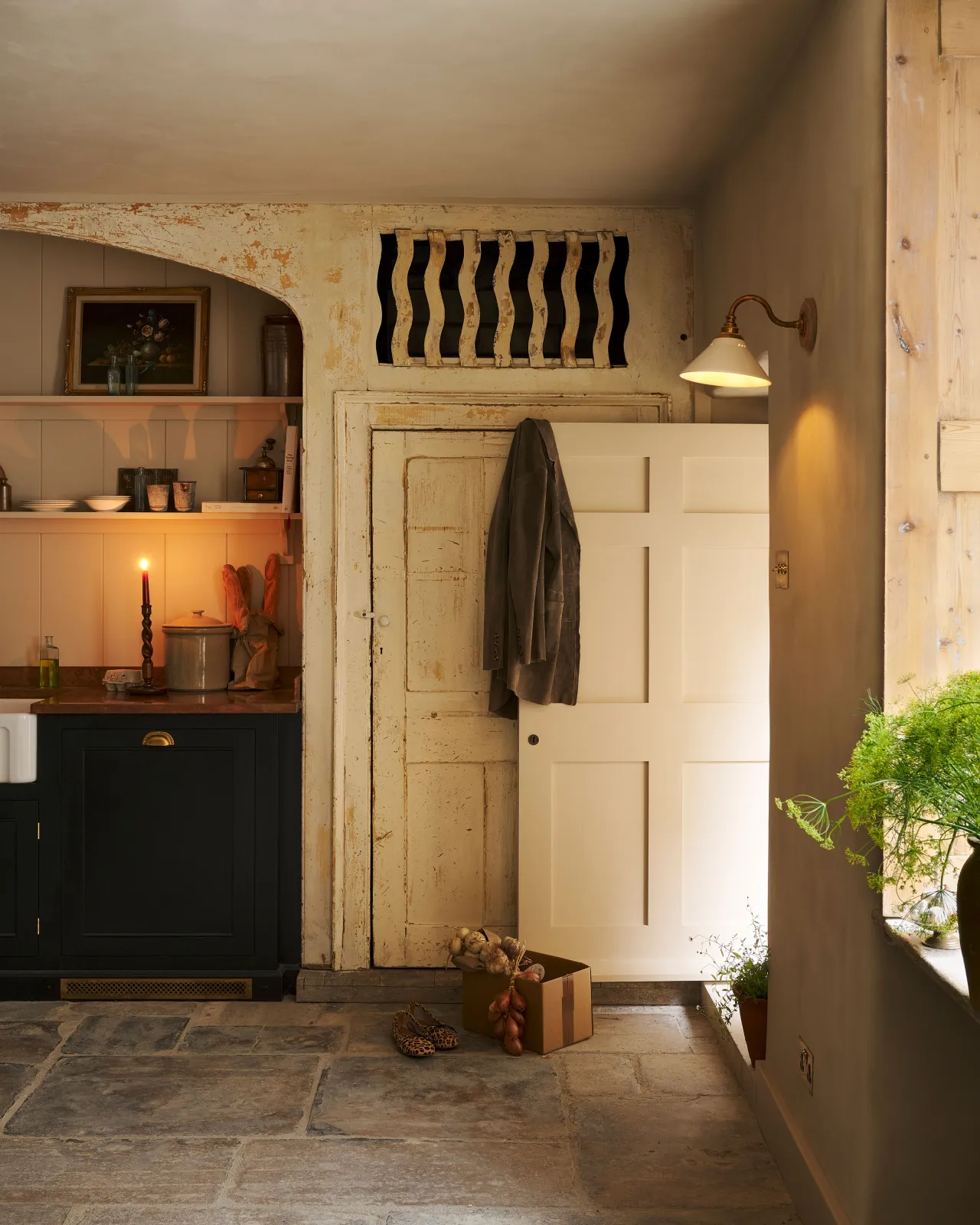 A rustic kitchen with black painted Georgian-style cabinetry, stone flooring and crumbling cream painted walls and joinery. A small Victorian-inspired wall light with a swan neck brass arm and a cream glazed ceramic shade is fixed to the wall by an open door.