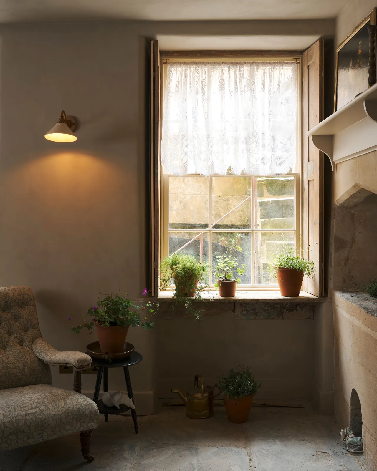 Light pours through a large window into a dark room with a small Victorian-inspired wall light with a swan neck brass arm and a cream glazed ceramic shade is fixed to the wall by the window.
