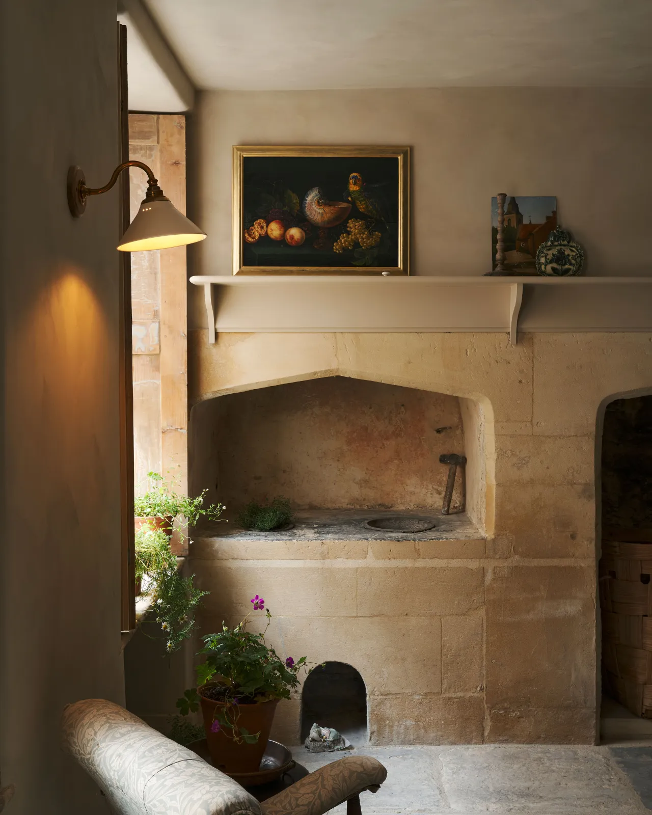 An atmospheric room with a fireplace built into a stone wall by a tall window. A small Victorian-inspired wall light with a swan neck brass arm and a cream glazed ceramic shade is fixed to the wall by the window.
