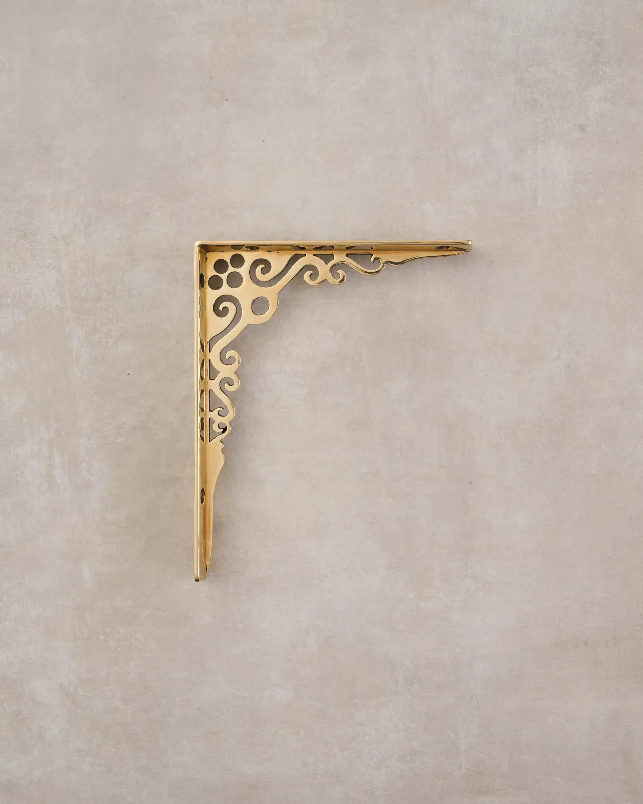 A side profile view of a small Victorian-inspired ornate shelf bracket in a warm polished brass finish.
