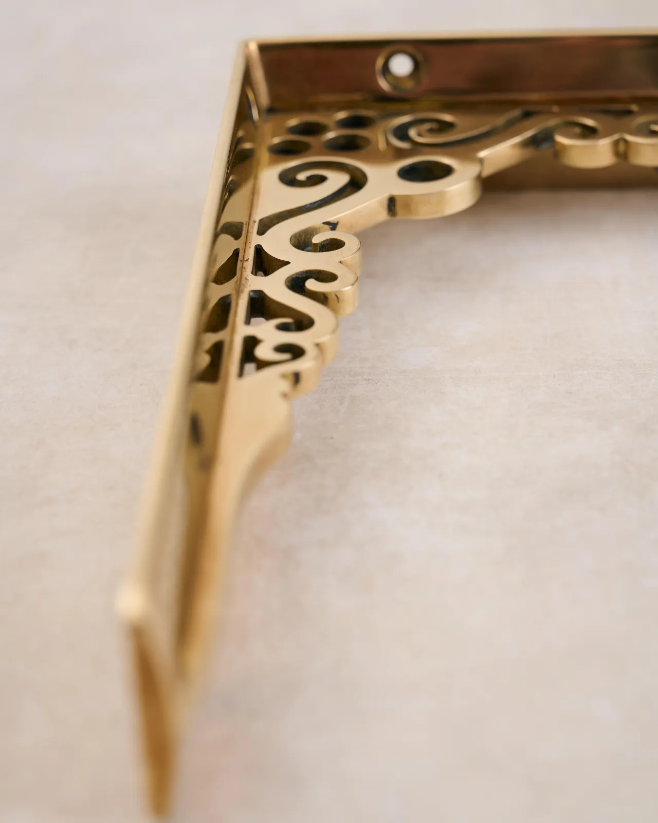 A close-up view of the decorative cutout patterns on a small Victorian-inspired ornate polished brass shelf bracket.