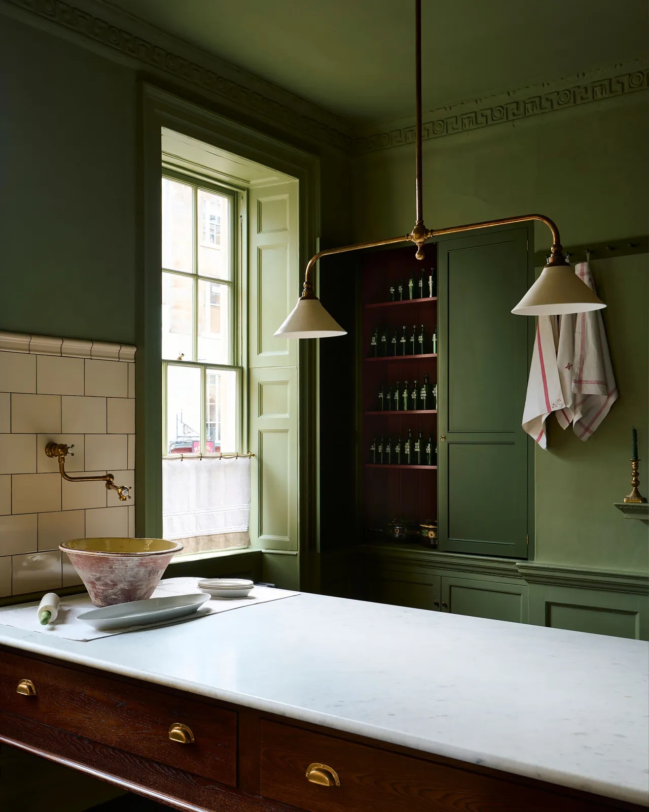 An oblique view of a Victorian-inspired chandelier light with brass arms and small unglazed porcelain shades in a dark green room with a large oak prep table with a Carrara marble worktop. The light is off.