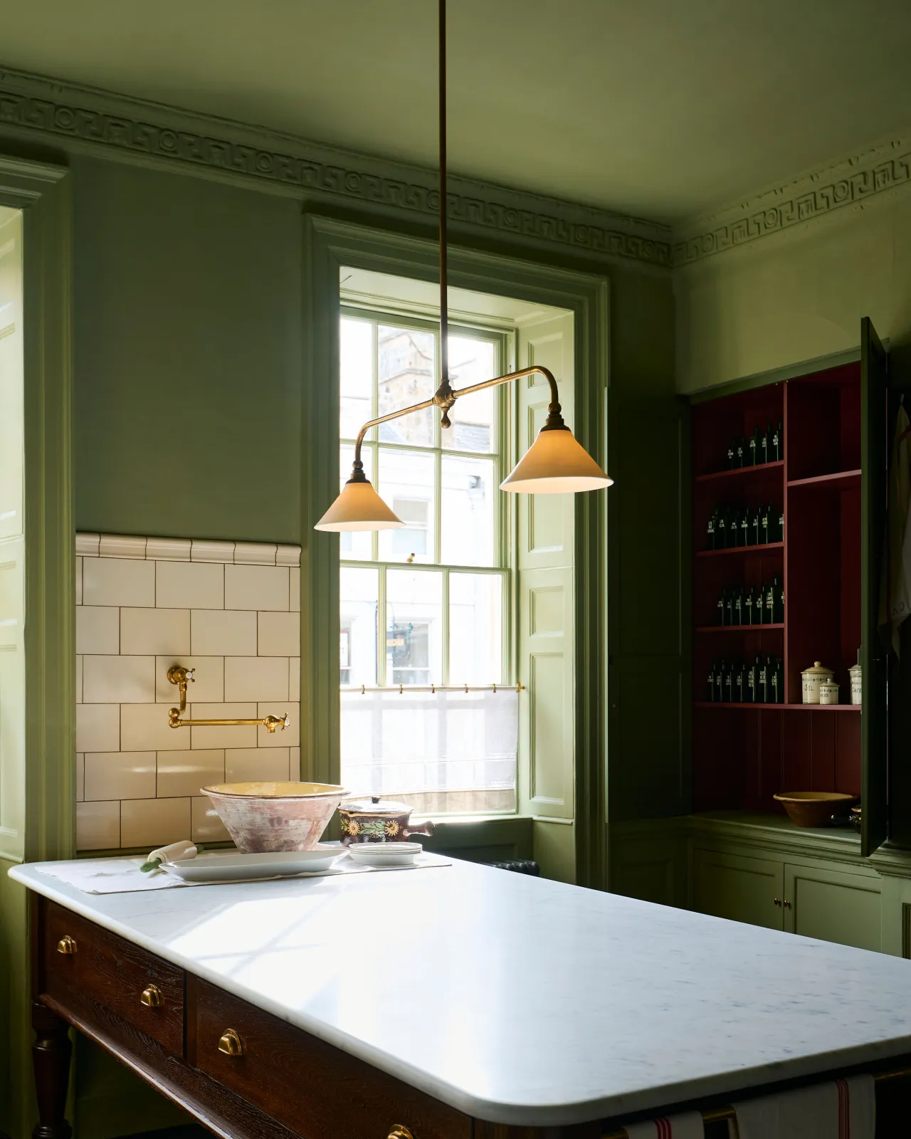 An oblique view of a Victorian-inspired chandelier light with brass arms and small unglazed porcelain shades in a dark green room with a large oak prep table with a Carrara marble worktop. The light is on, showing the warm red glow of the translucent surface.
