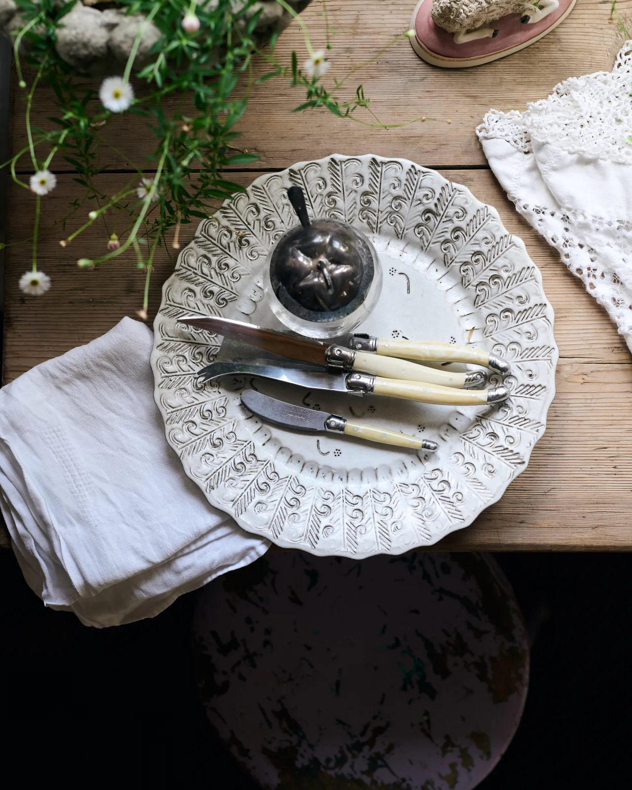 An overhead view of a round handmade bohemian-style white ceramic platter with delicate impressions and surface patterns, complimented perfectly by vintage silverware, white linens and a rustic pine tabletop.