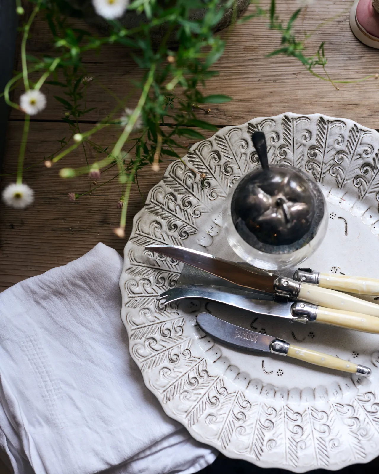 A close-up overhead view of a round handmade bohemian-style white ceramic platter with delicate impressions and surface patterns, complimented perfectly by vintage silverware, white linens and a rustic pine tabletop.