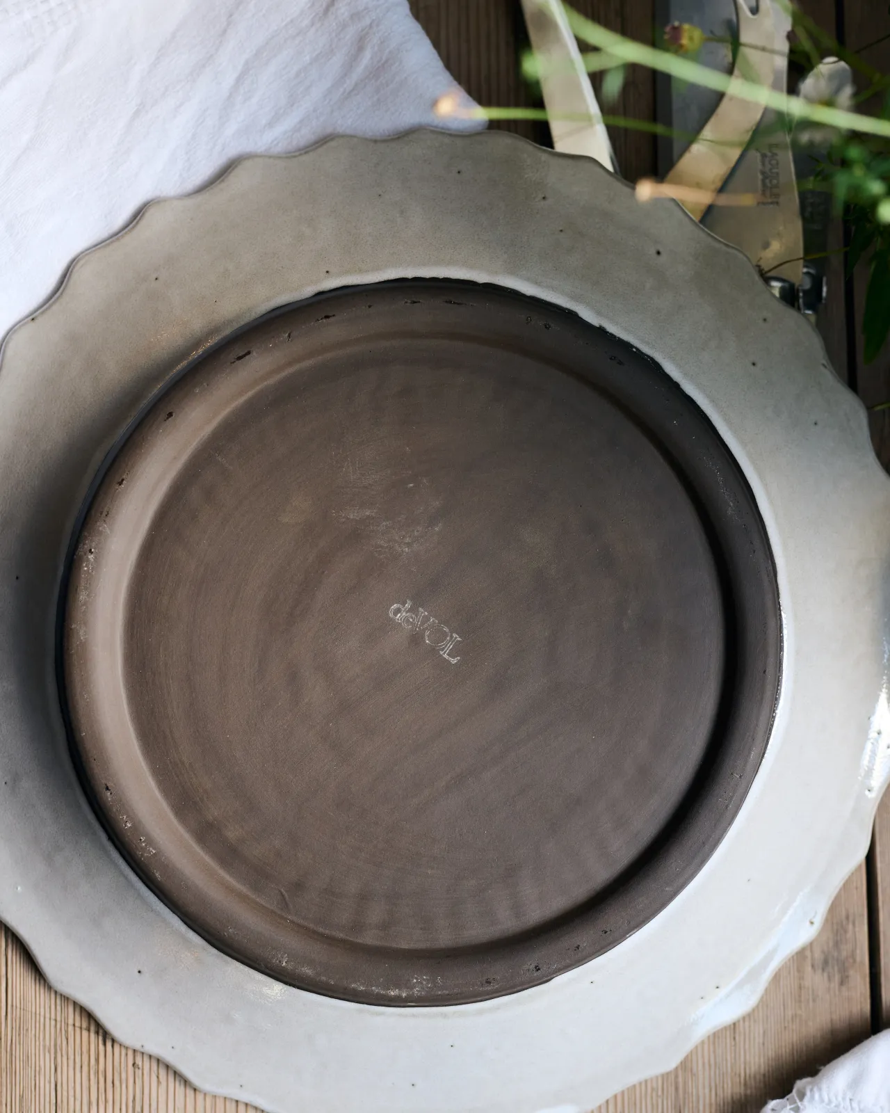 The underside of a round handmade bohemian-style ceramic platter. A white glazed rim surrounds an unglazed chocolate-coloured clay foot with a 'deVOL' logo in the centre.