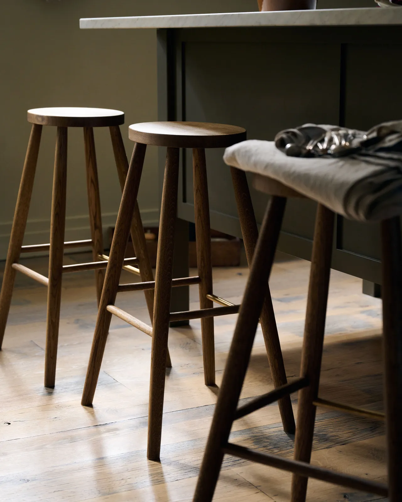 An oblique view of three simple oak bar stools with oval-shaped seats in front of an island breakfast bar.