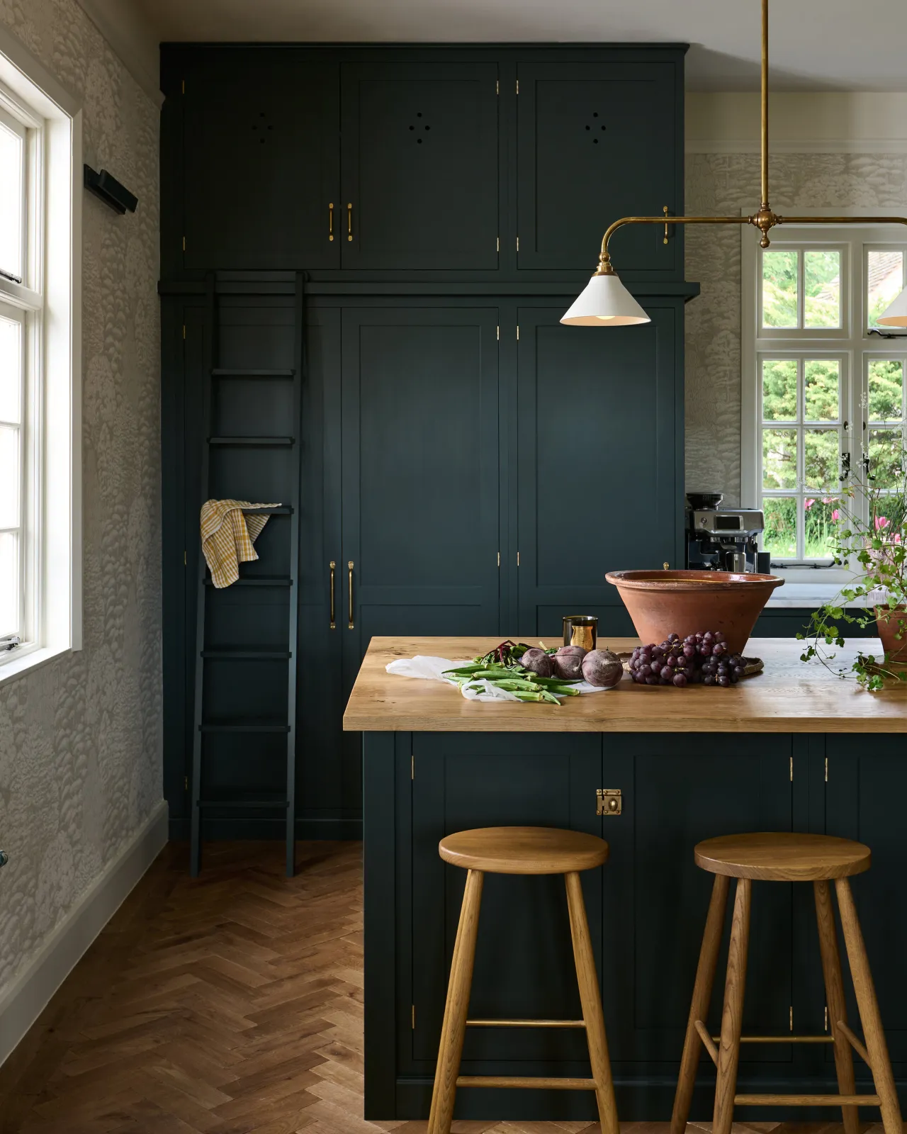 A straight-on view of a dark green painted Shaker-style kitchen with an island breakfast bar and two simple oak bar stools with oval-shaped seats and brass footrails.