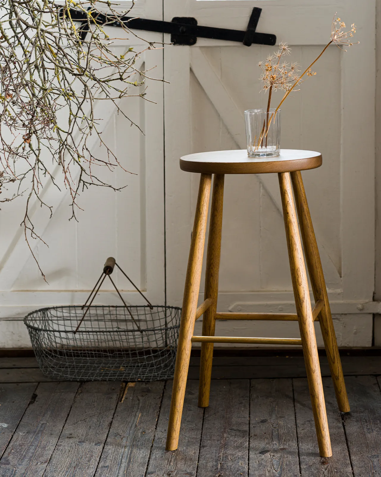 A straight-on view of a simple oak bar stool with an oval-shaped seat and a brass footrail with dried flowers in a glass on top.