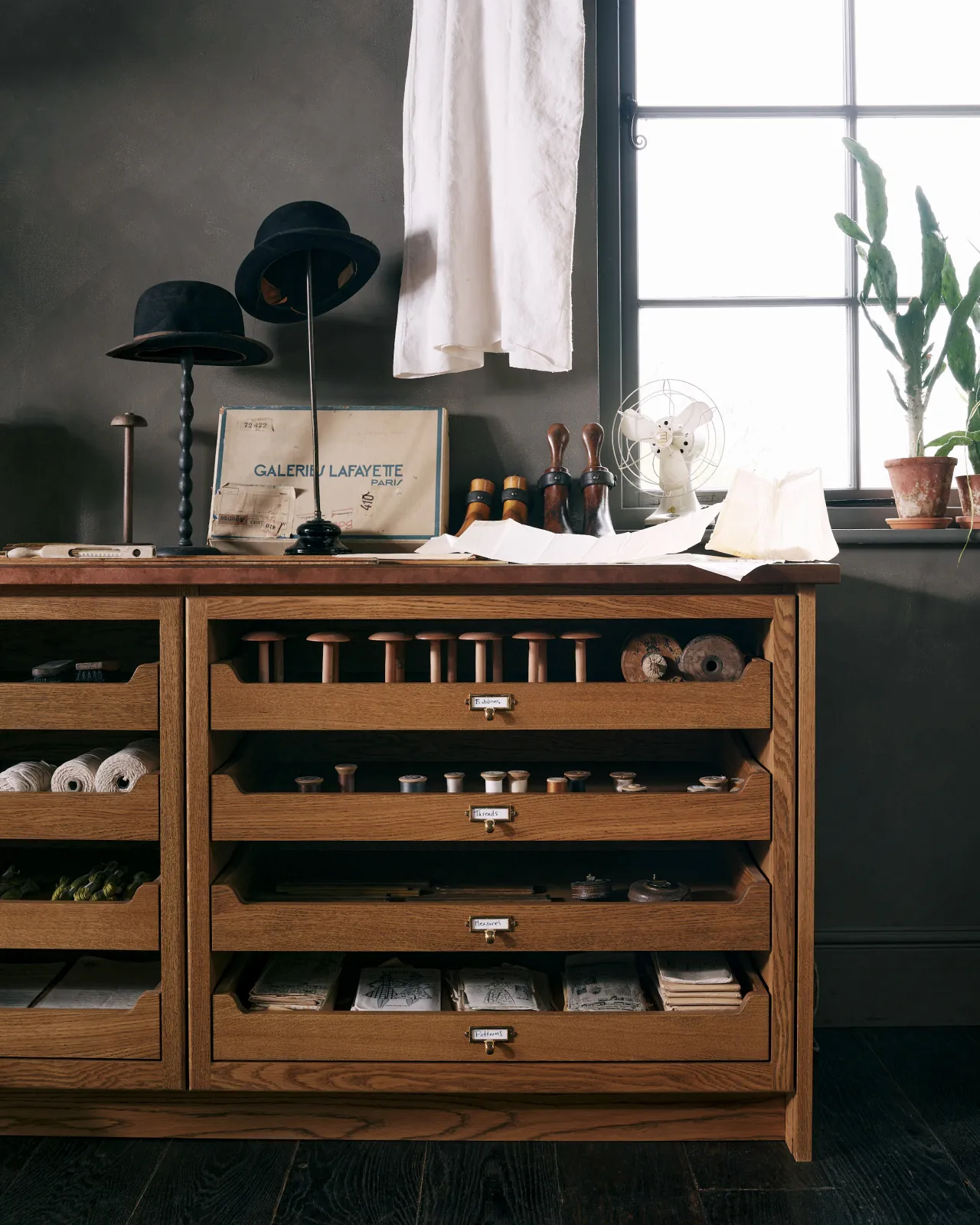 A straight-on view of an oak mid-century-style cupboard with partially open drawers in a dark room with vintage styling. Each drawer has a small cardframe pull handle in a warm burnished brass finish and handwritten paper label.
