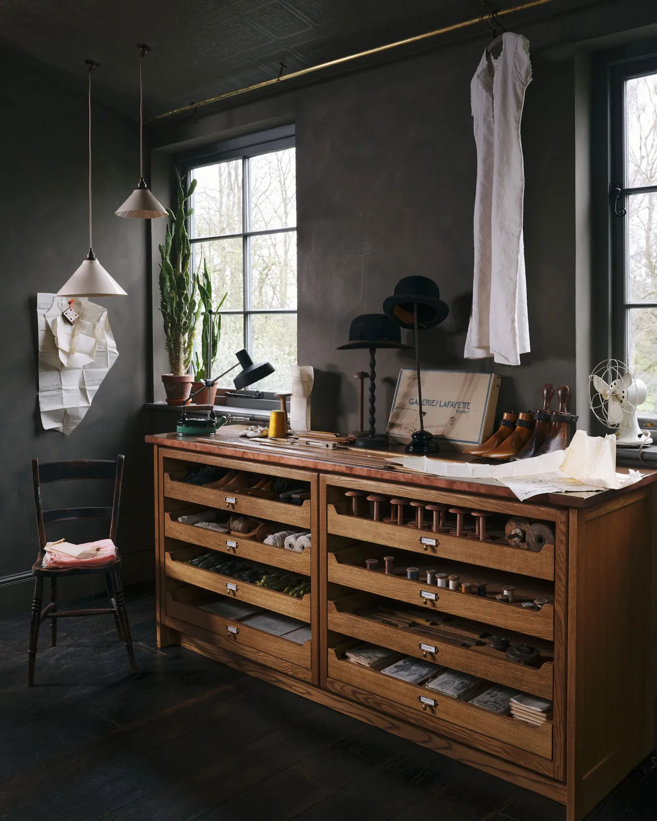 An oblique view of an oak mid-century-style cupboard with partially open drawers in a dark room with vintage styling. Each drawer has a small cardframe pull handle in a warm burnished brass finish and handwritten paper label.