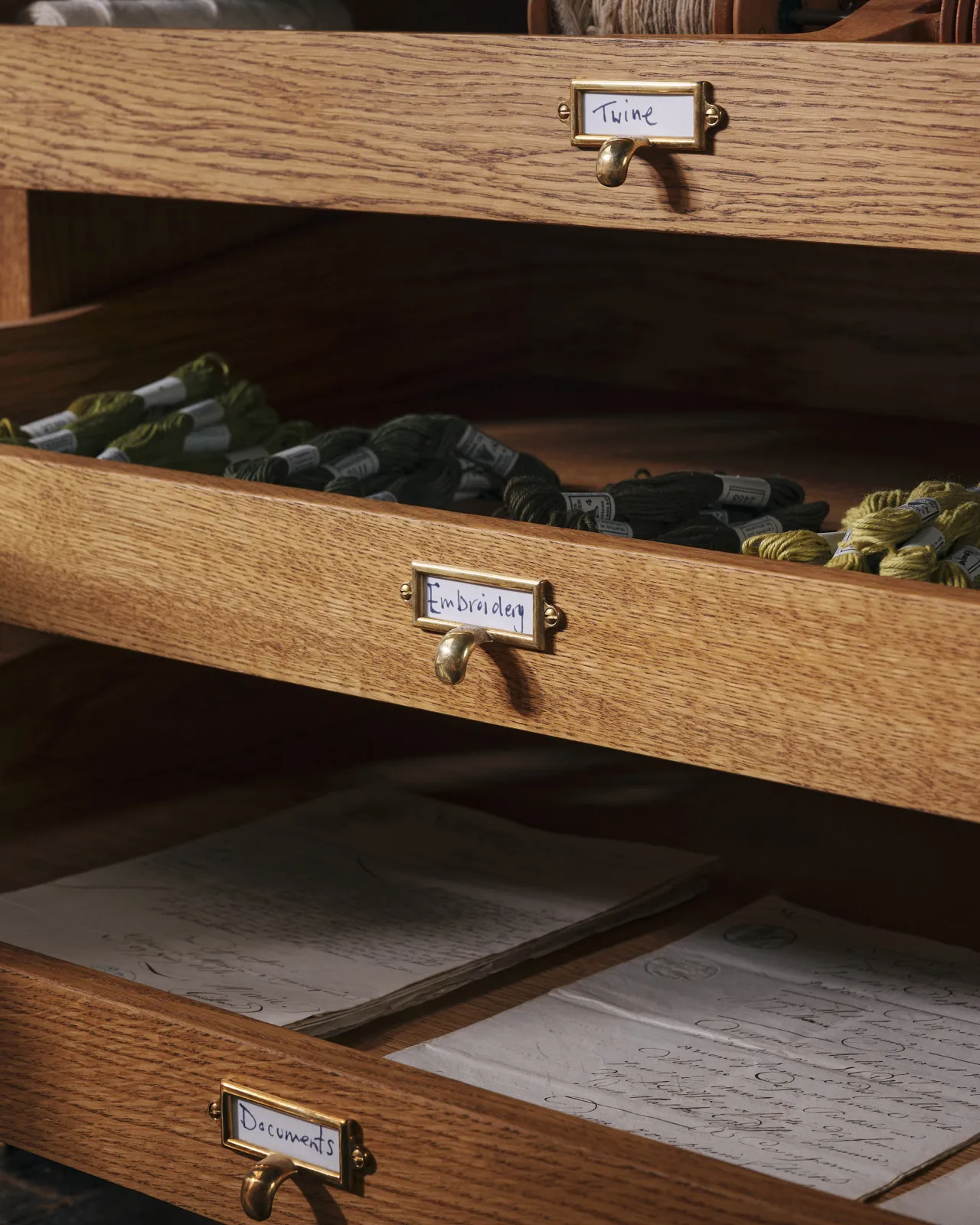 An oblique view of an oak mid-century-style cupboard with partially open drawers, fitted with small cardframe pull handles in a warm burnished brass finish and handwritten paper labels that read 'Twine', 'Embroidery', and 'Documents'.