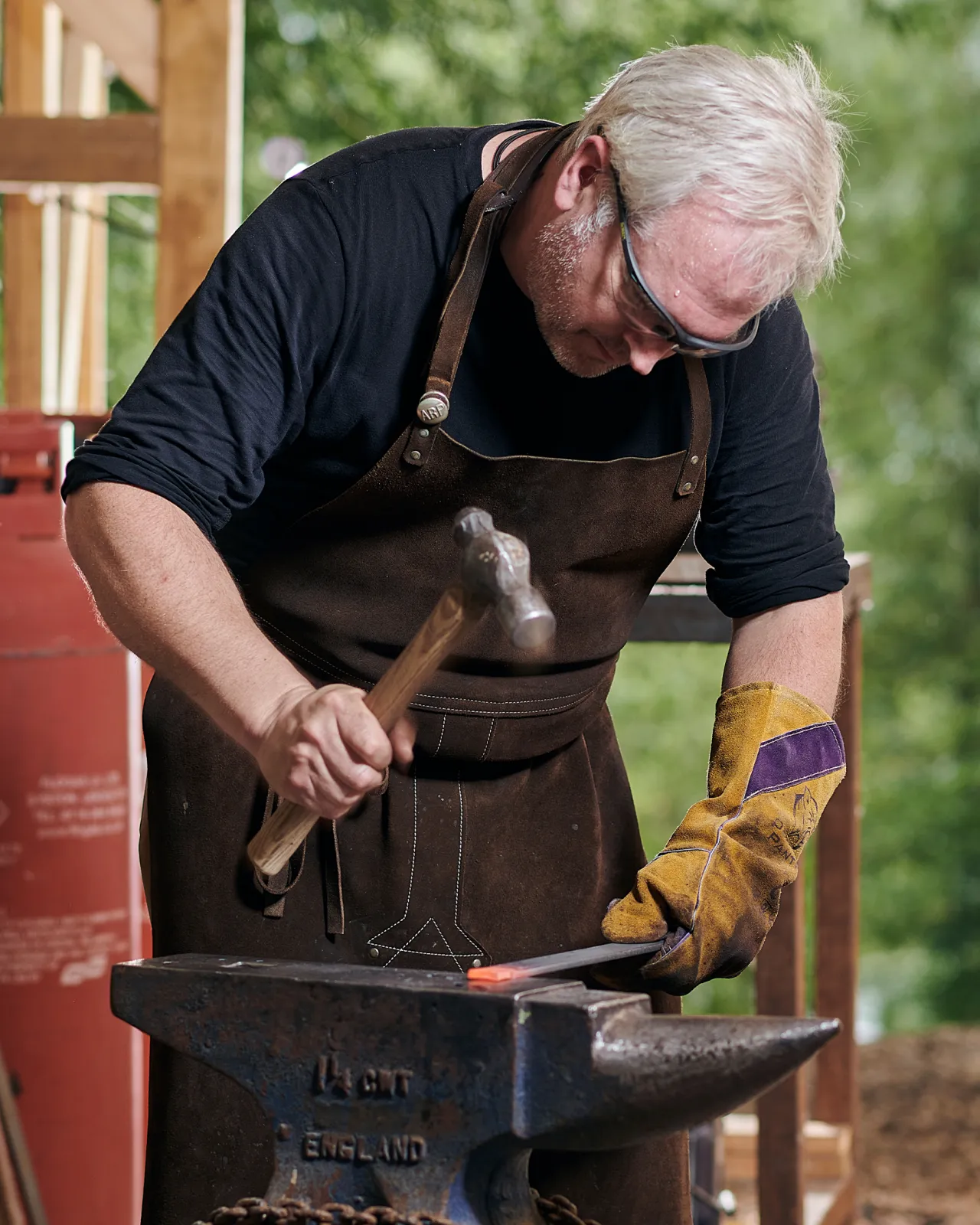 A blacksmith in a brown apron is hammering a piece of red-hot iron on an anvil.
