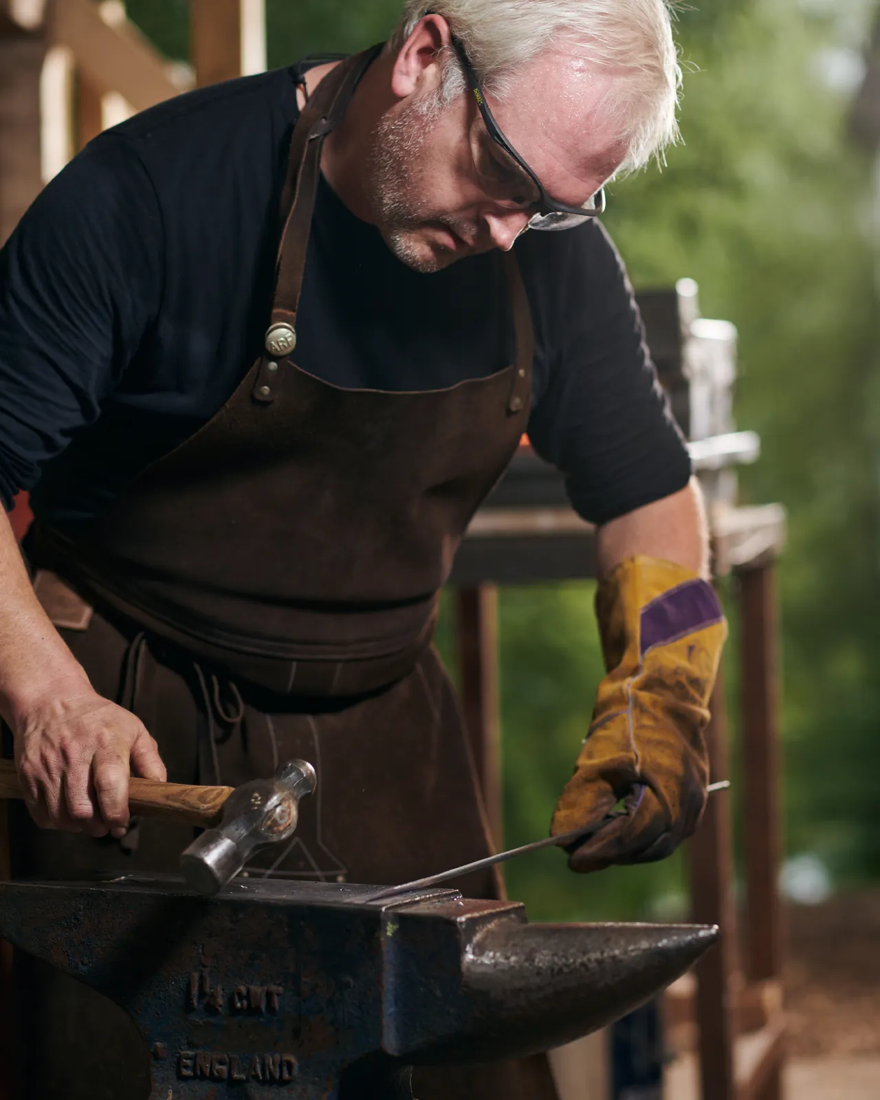A blacksmith in a brown apron is hammering the end of an iron rod into a sharp point on an anvil.